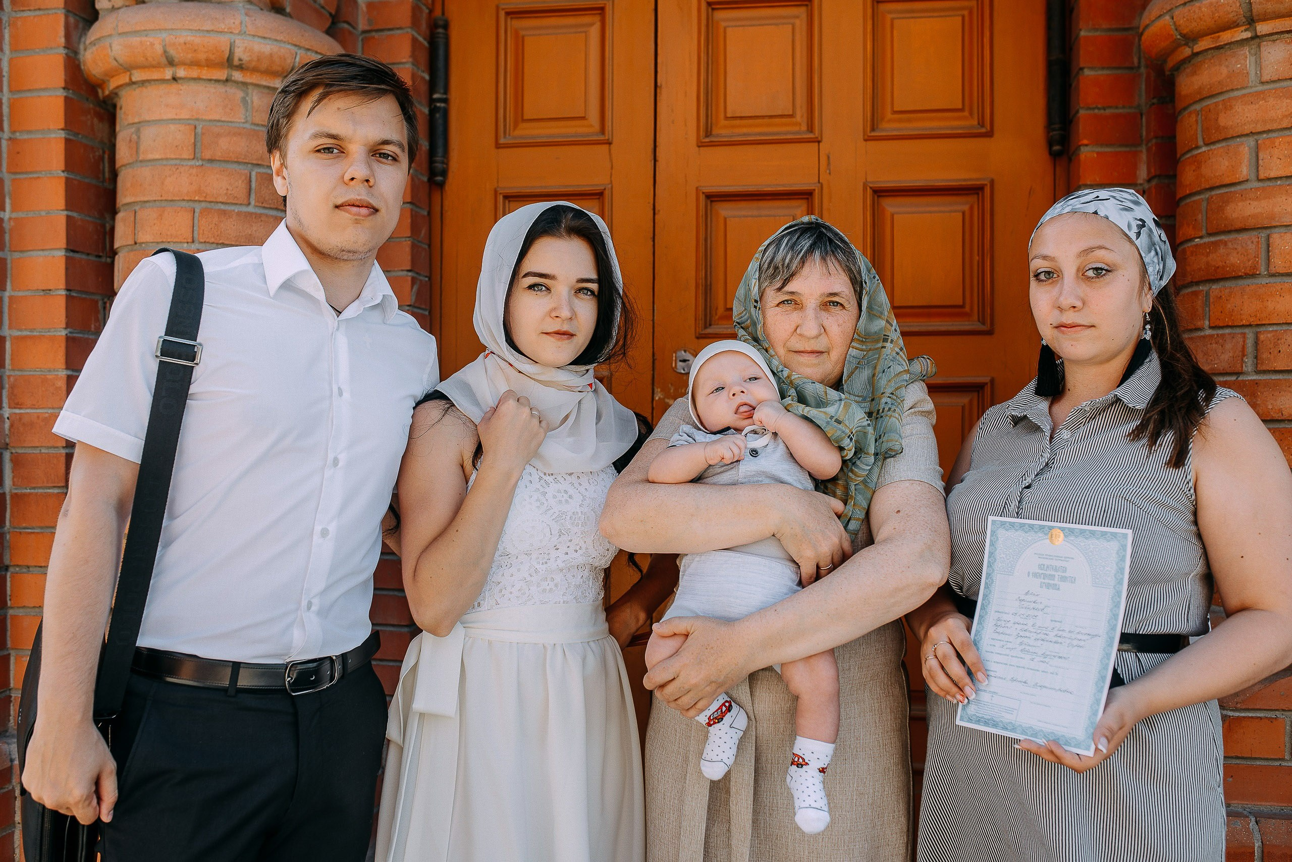 Семейное фото после крестин: родители и крестные с ребенком и свидетельством о крещении у входа в церковь.