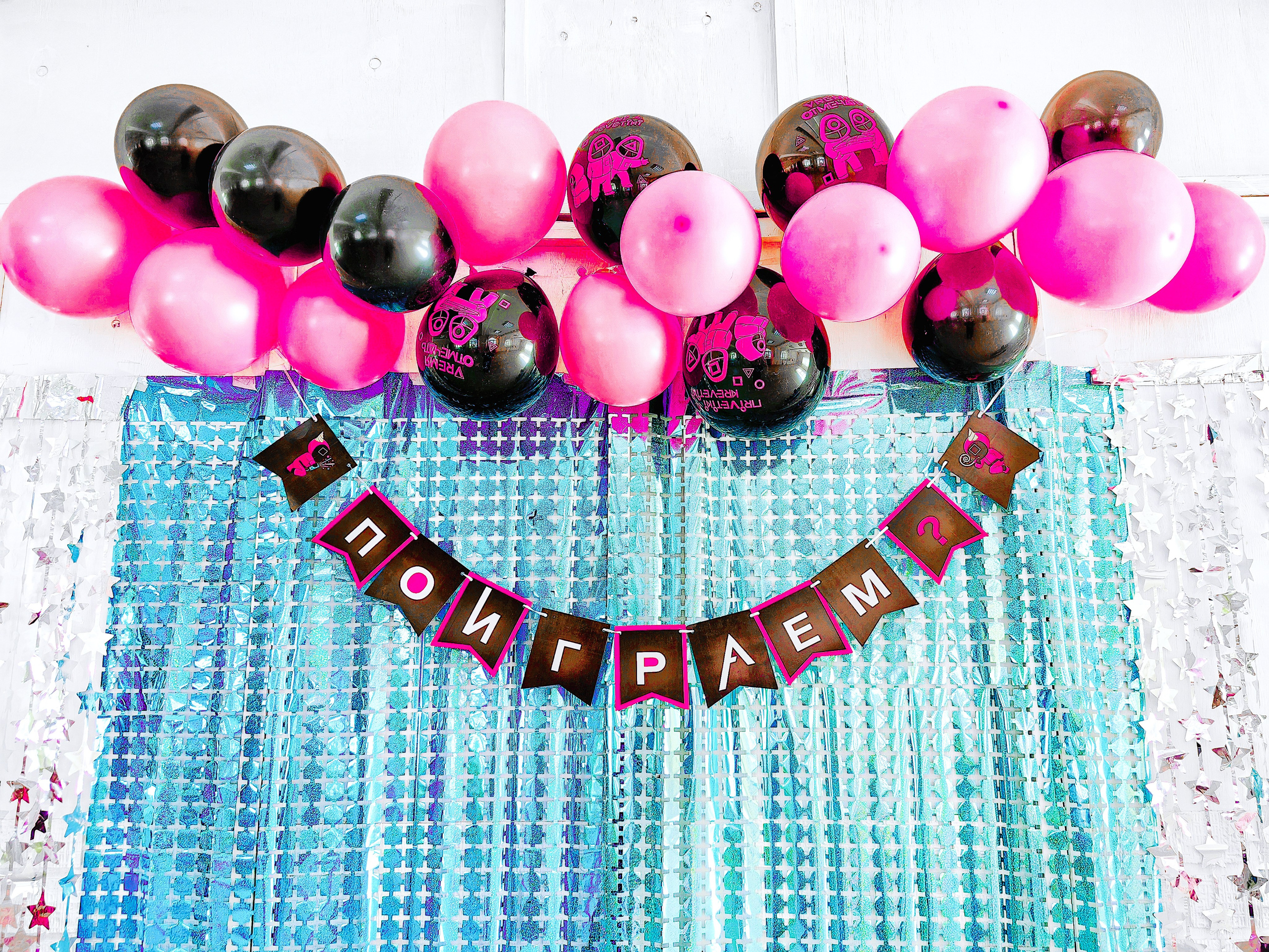 Вывеска "Поиграем" в черно-желтых тонах расположена на стене пож пучками розово-черных шаров на бирюзовом фоне - фрагмент фотозоны для дня рождения в стиле Игры в кальмара