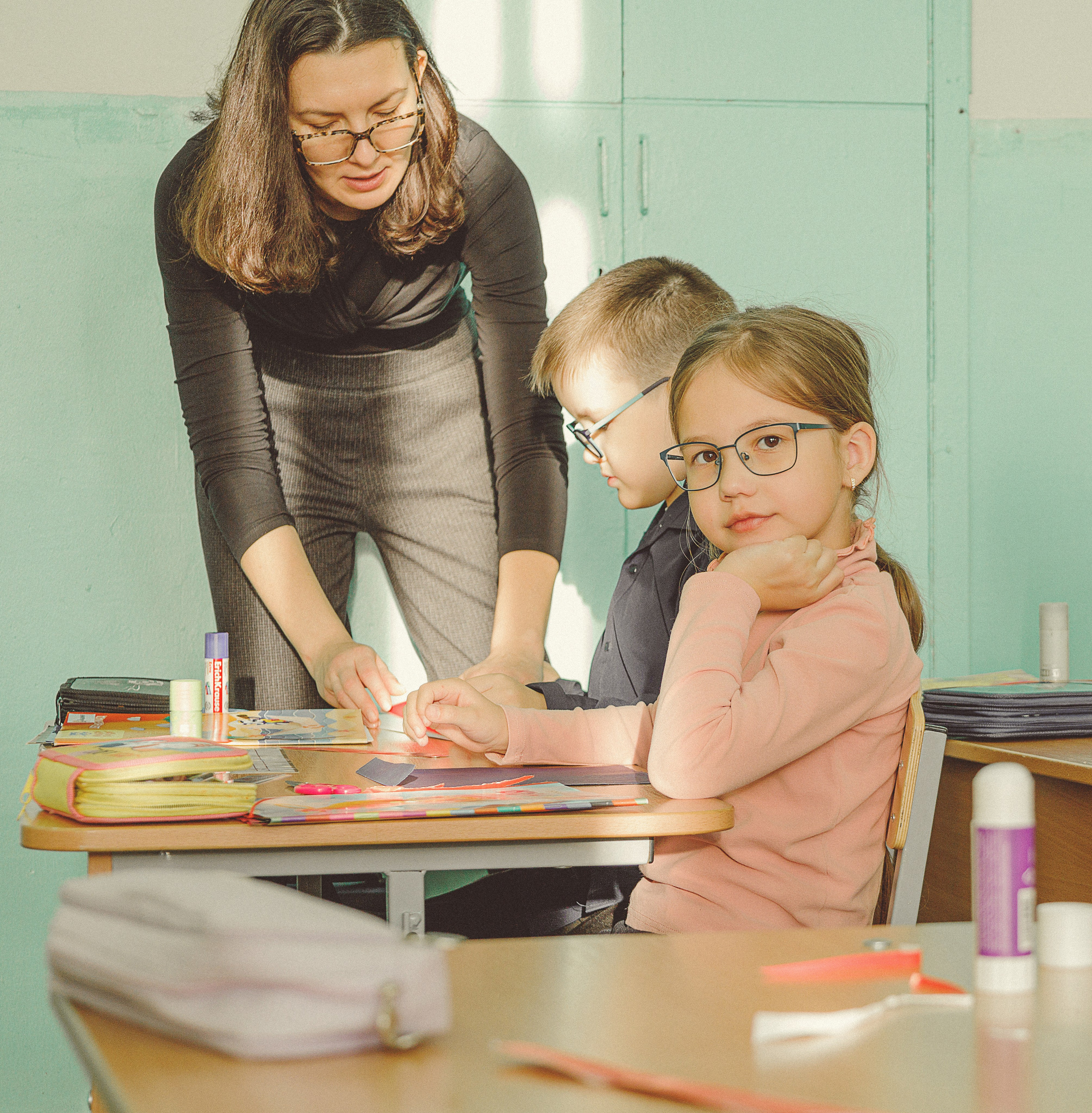 Учительница помогает ребенку с поделкой за письменным столом в классе. Четверо внимательных детей работают над красочными проектами. Свет из окна добавляет тепла.