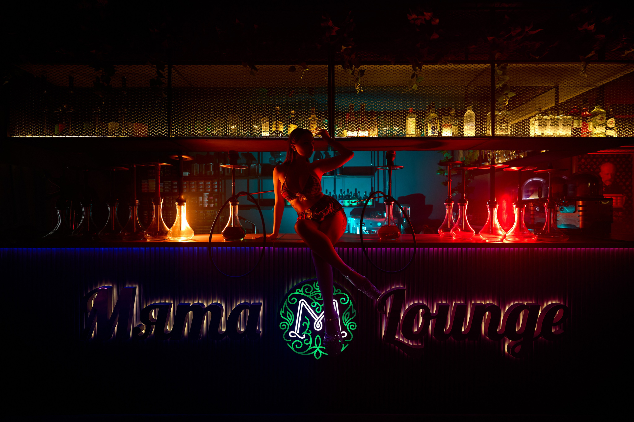 Фотосъёмка контента для lounge бара 'Мята'. На фотографии — женщина, позирующая на барной стойке в ярко освещённом пространстве. Бар оформлен в стильных и современных оттенках с подсветкой, создающей атмосферу вечеринки. На заднем плане видны бутылки с алкоголем, а над барной стойкой ярко светится логотип заведения. Окружение погружает в расслабленную и элегантную атмосферу ночного клуба