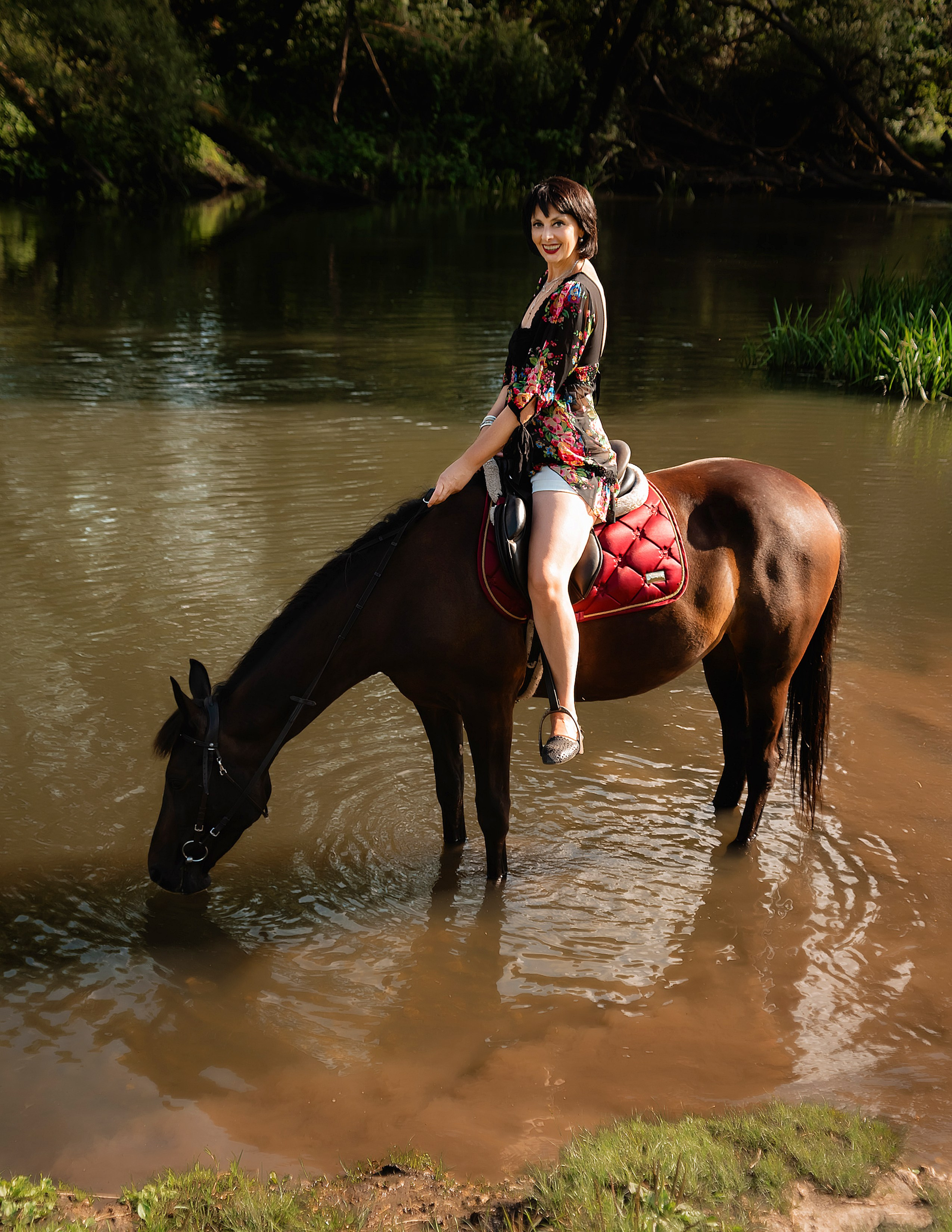 На фото женщина в бежевом платье стоит с лошадью в пруду.