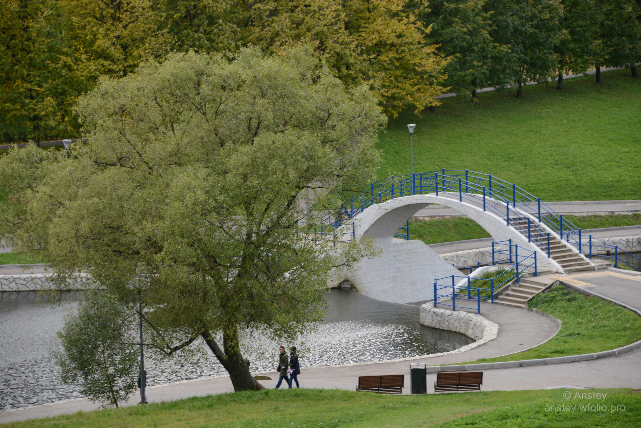 Парк Олимпийской деревни в Москве ранней осенью. Пешеходный мост через протоку между Олимпийскими прудами. Pond, bridge, trees and buildings in city against sky. Walk in a Moscow city park in early autumn season.
