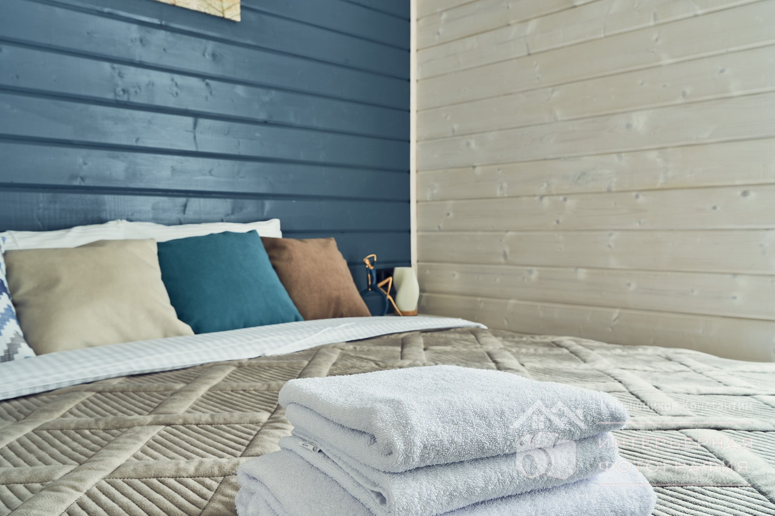 Интерьерная съемка пушистых полотенец на переднем плане и подушек на двуспальной кровати в загородном доме