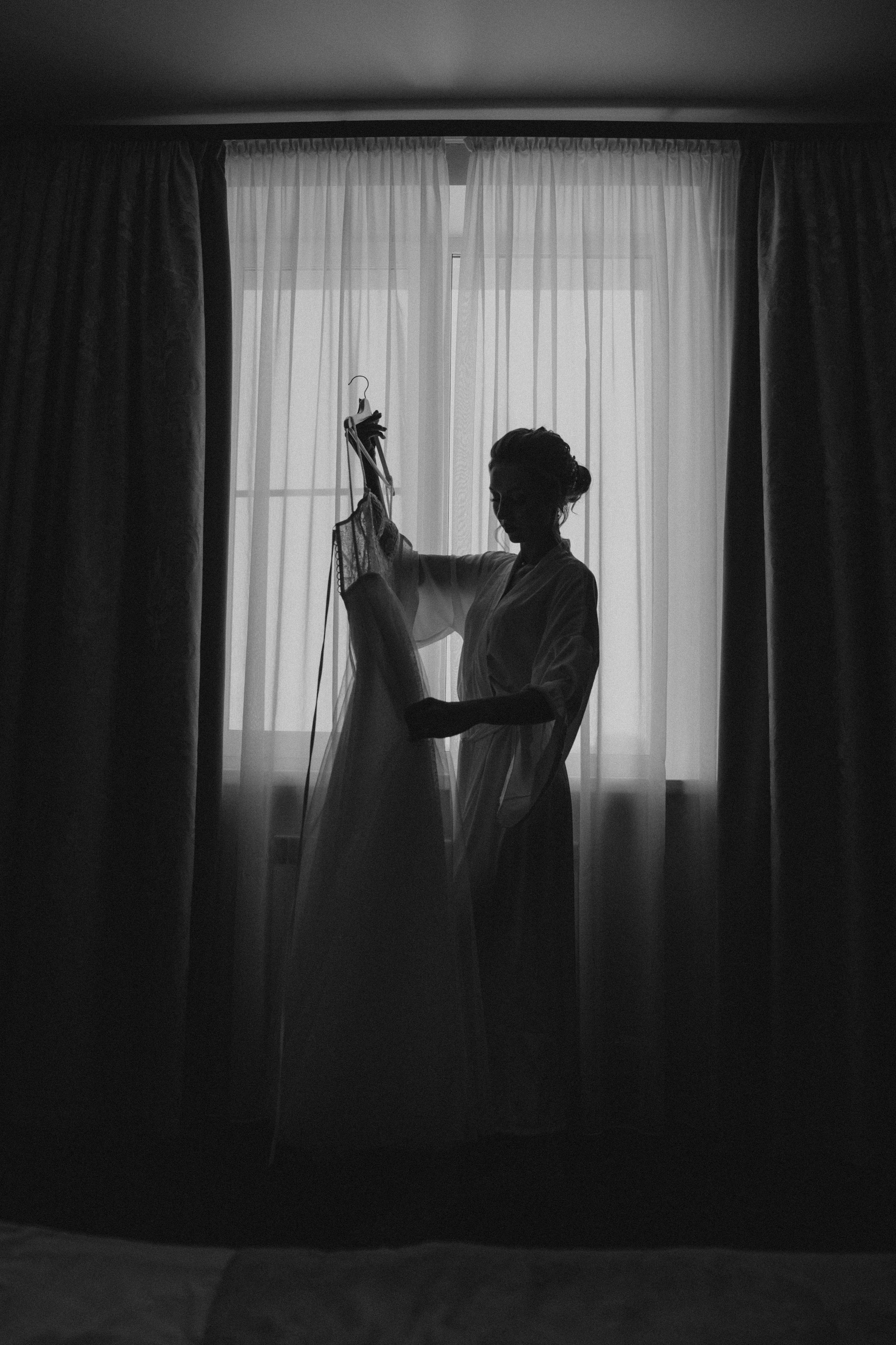 Невеста рассматривает платье на фоне окна, силуэтное черно-белое фото