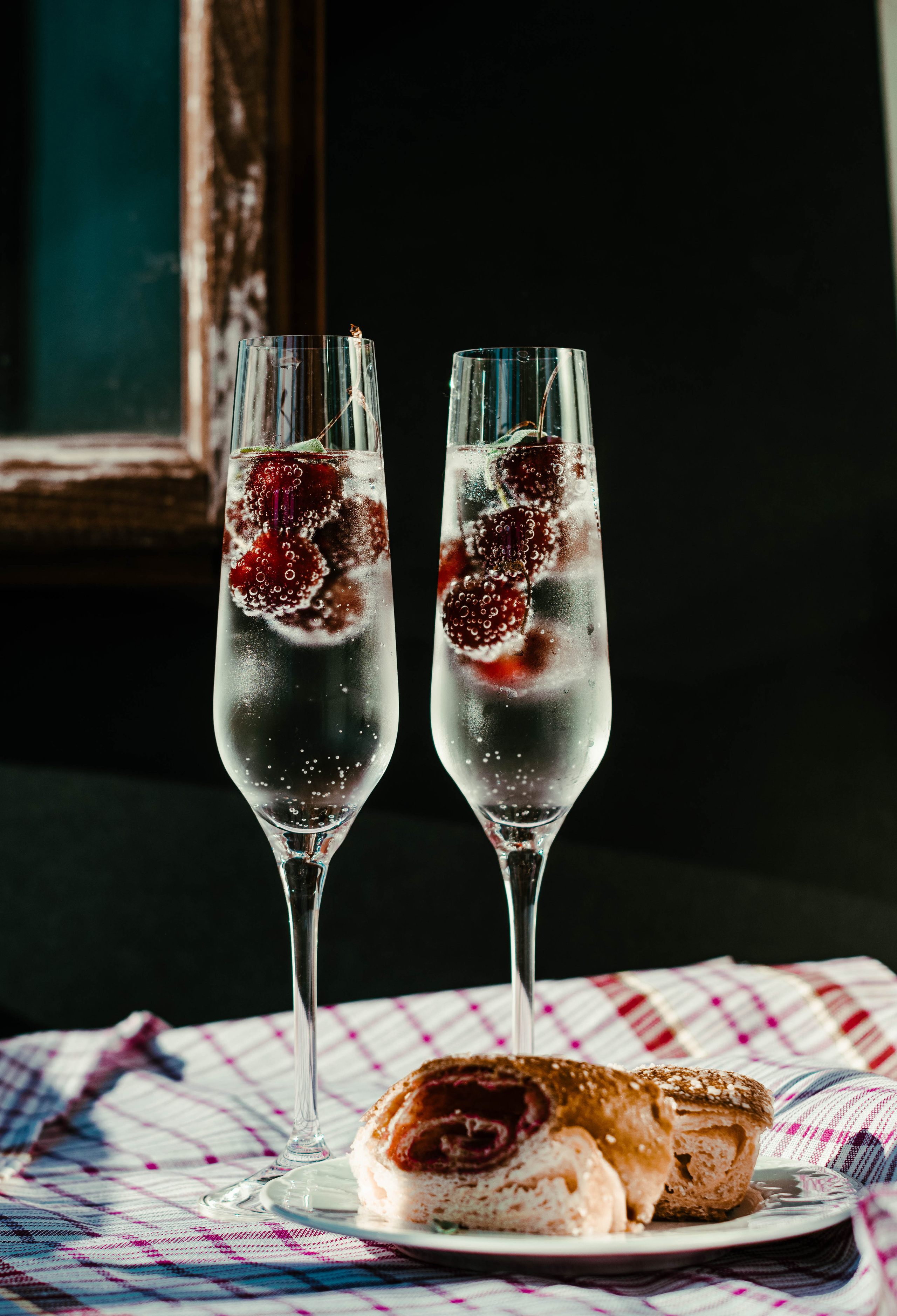 Два бокала с газированным напитком, вишней и тарелка с десертом в лучах солнца.