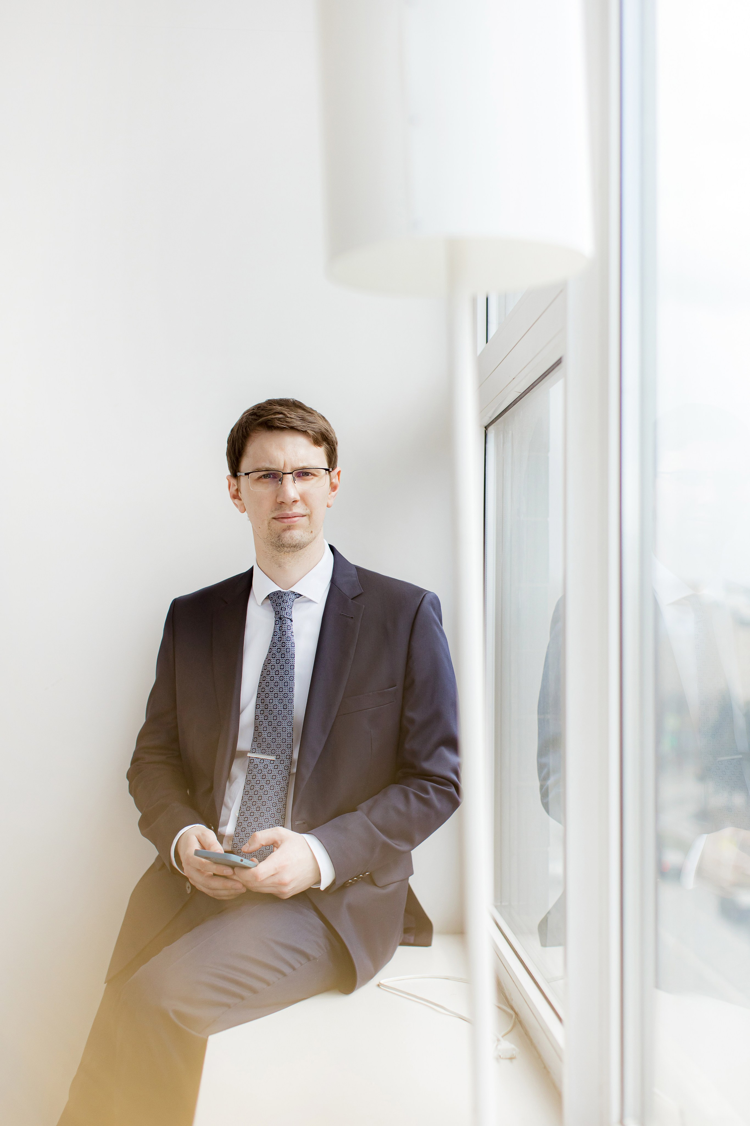 Мужчина в костюме с галстуком сидит у окна с телефоном в руках