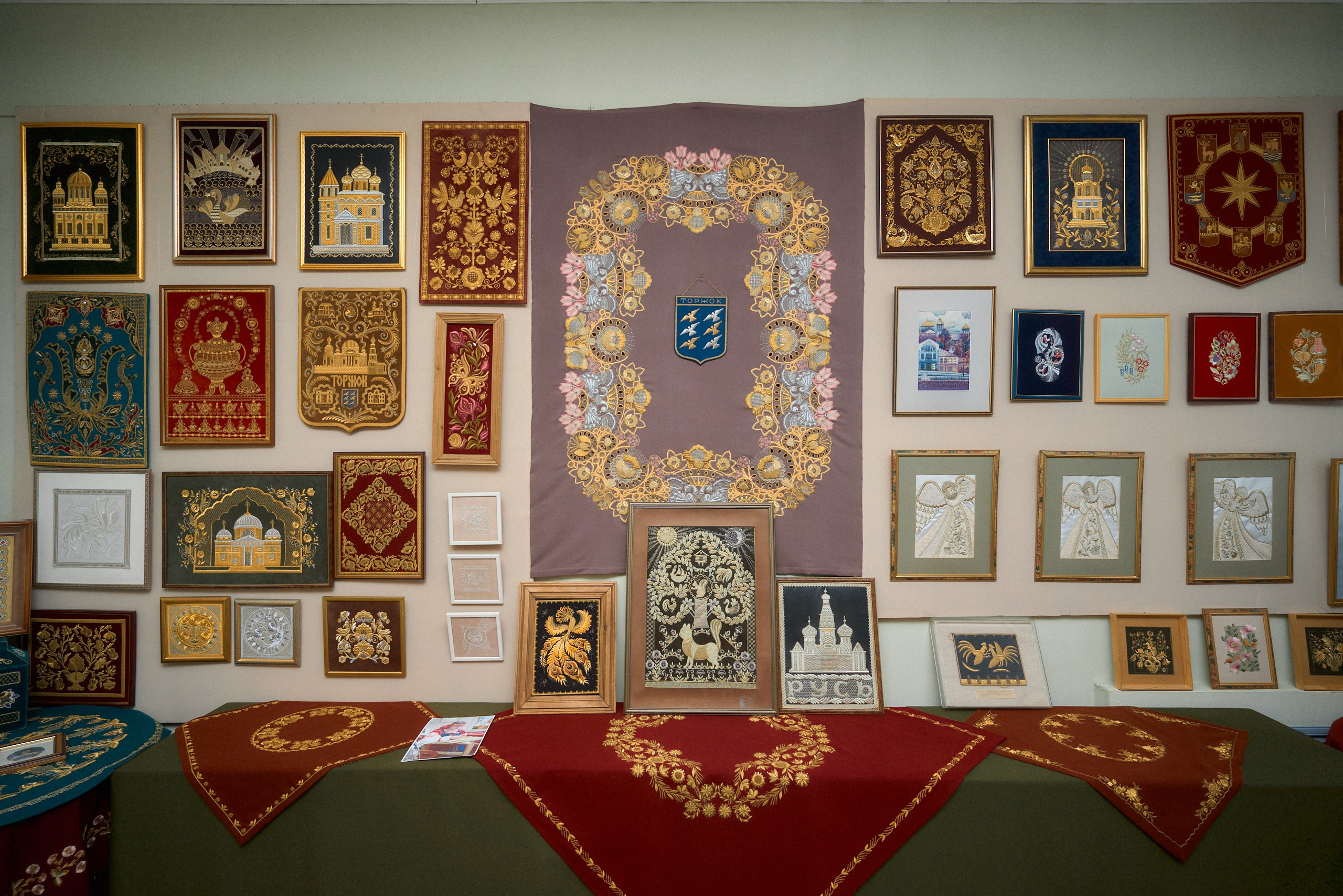 Альт текст для изображения: "Экспозиция в Музее торжокских золотошвей в Торжке, демонстрирующая коллекцию вышитых золотыми нитями изделий, включая картины, гербы, декоративные элементы и традиционные платки, расположенные на стенах и столе