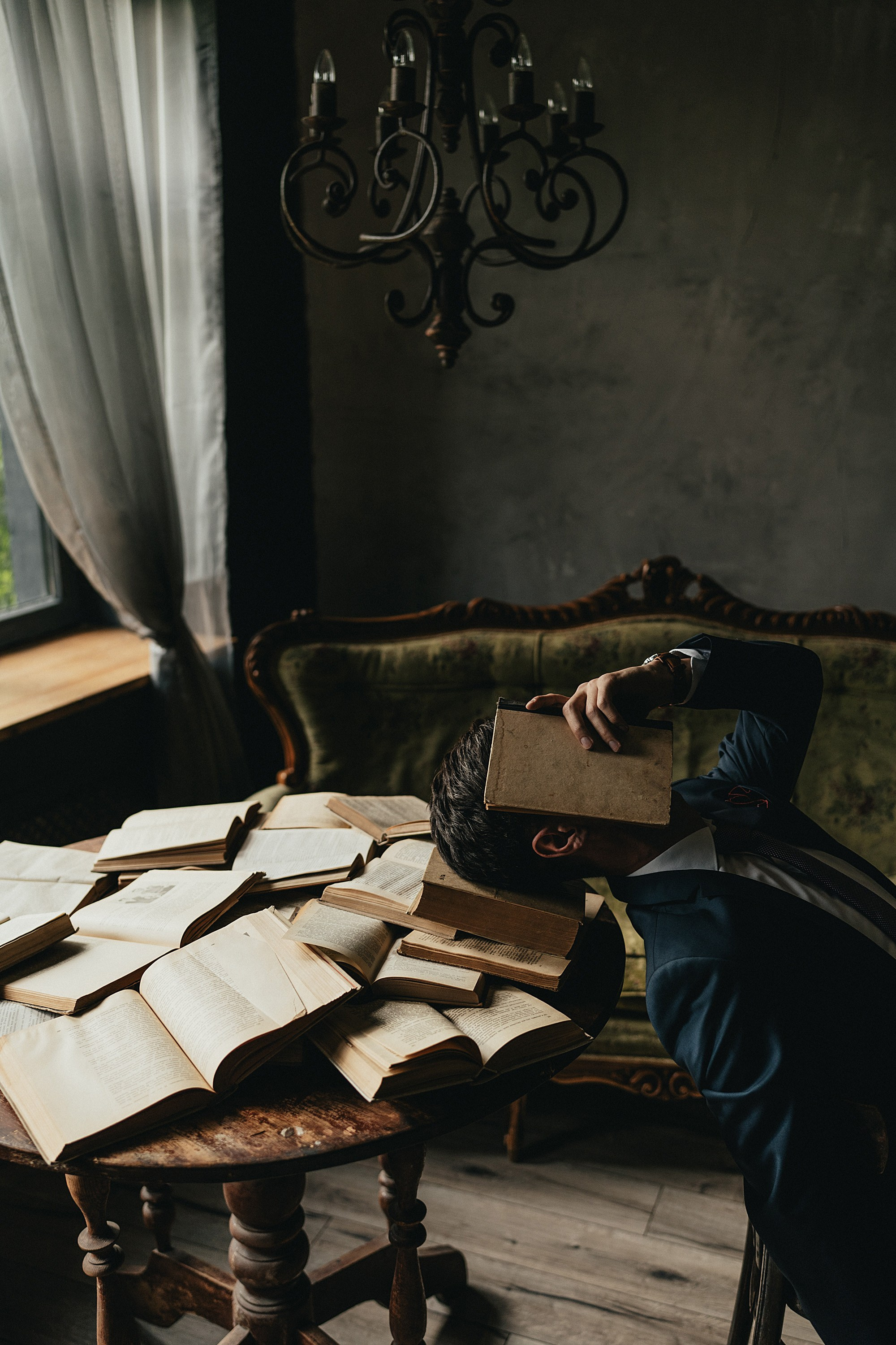 Мужчина в синем костюме закрывает лицо книгой, сидя за столом, уставленным книгами