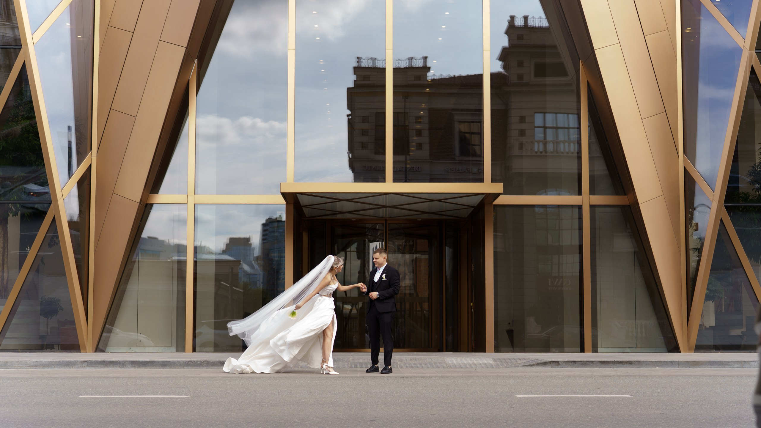 Молодожёны на фоне здания с зеркальными панелями и золотыми рамами: невеста в белом платье с длинным шлейфом, жених — в смокинге. Пара выглядит гармонично и счастливо | свадебный фотограф с опытом Екатеринбург