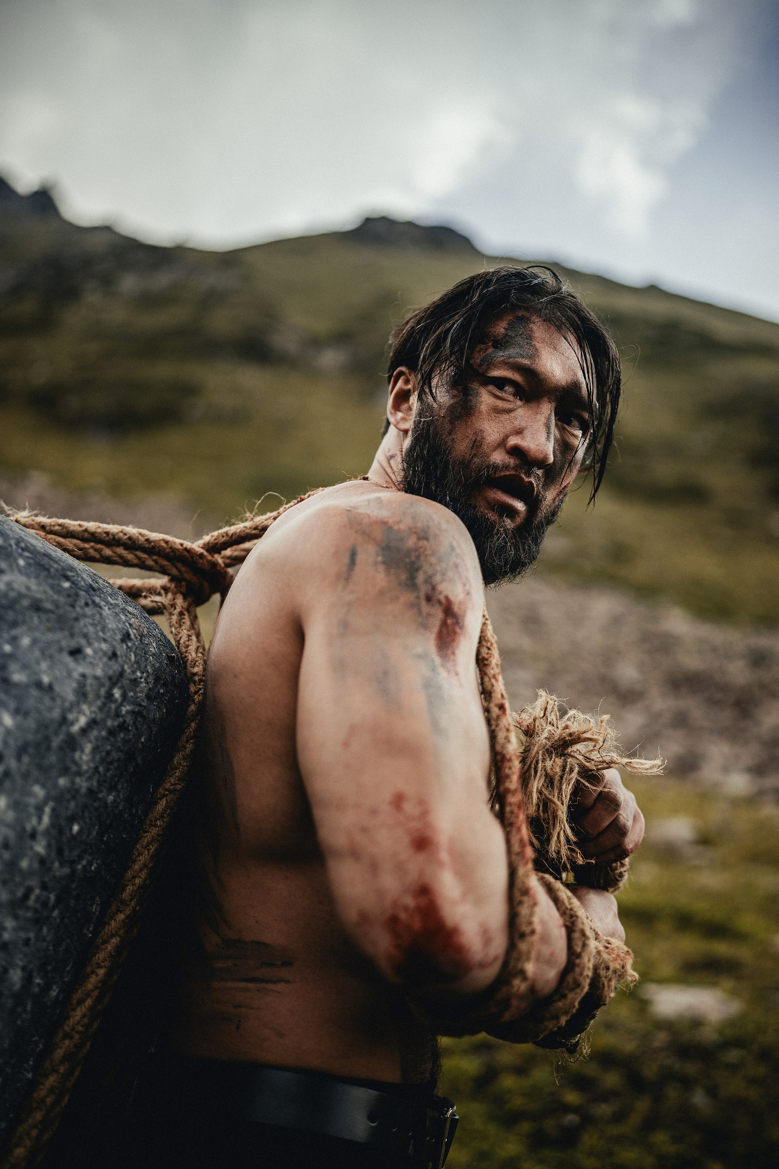 Мужчина с растрепанными волосами и бородой тянет веревку с камнем на горном фоне