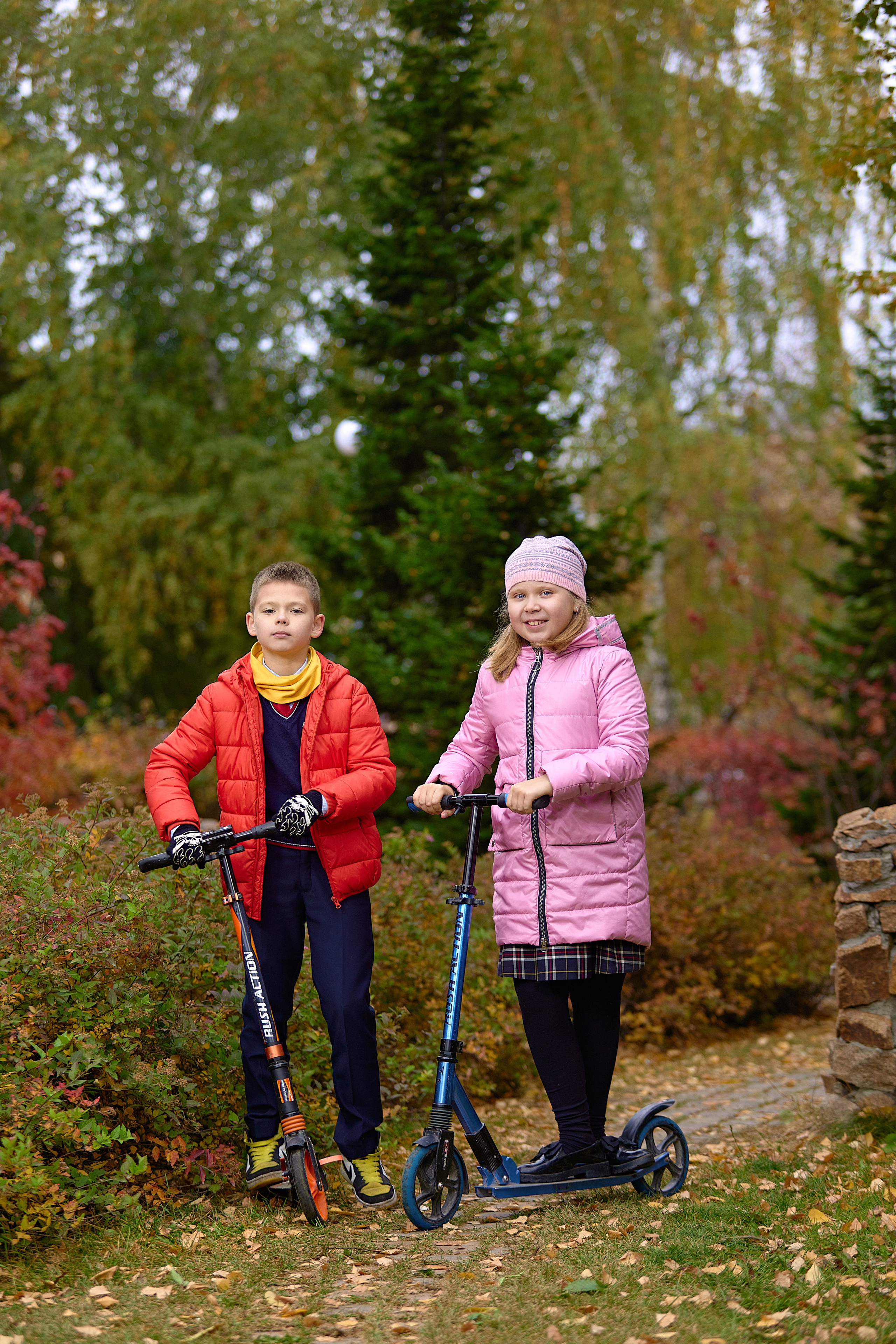 Мальчик и девочка стоят в осеннем парке и держат самокаты.