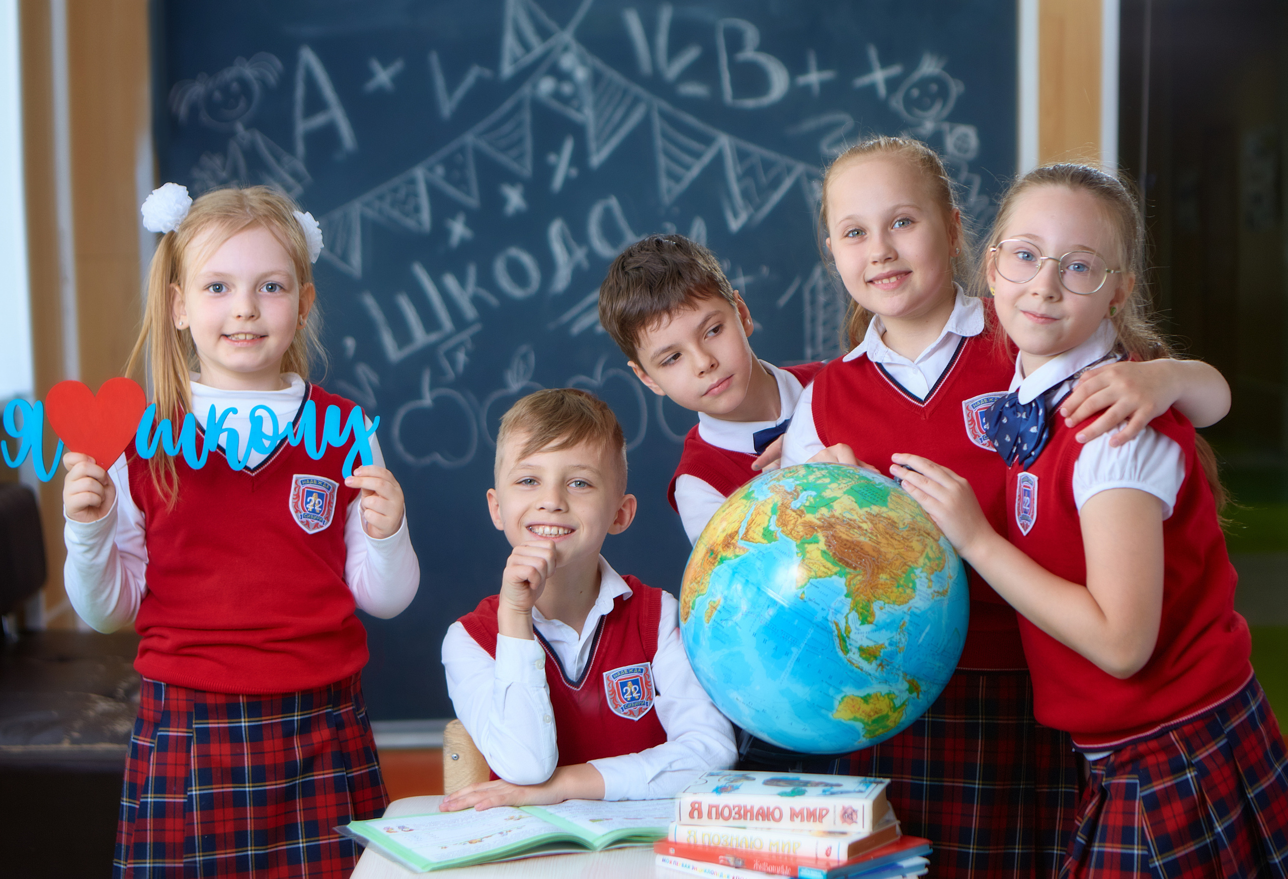 Первоклассники весело позируют фотографу с глобусом, надписью я люблю школу и учебником.