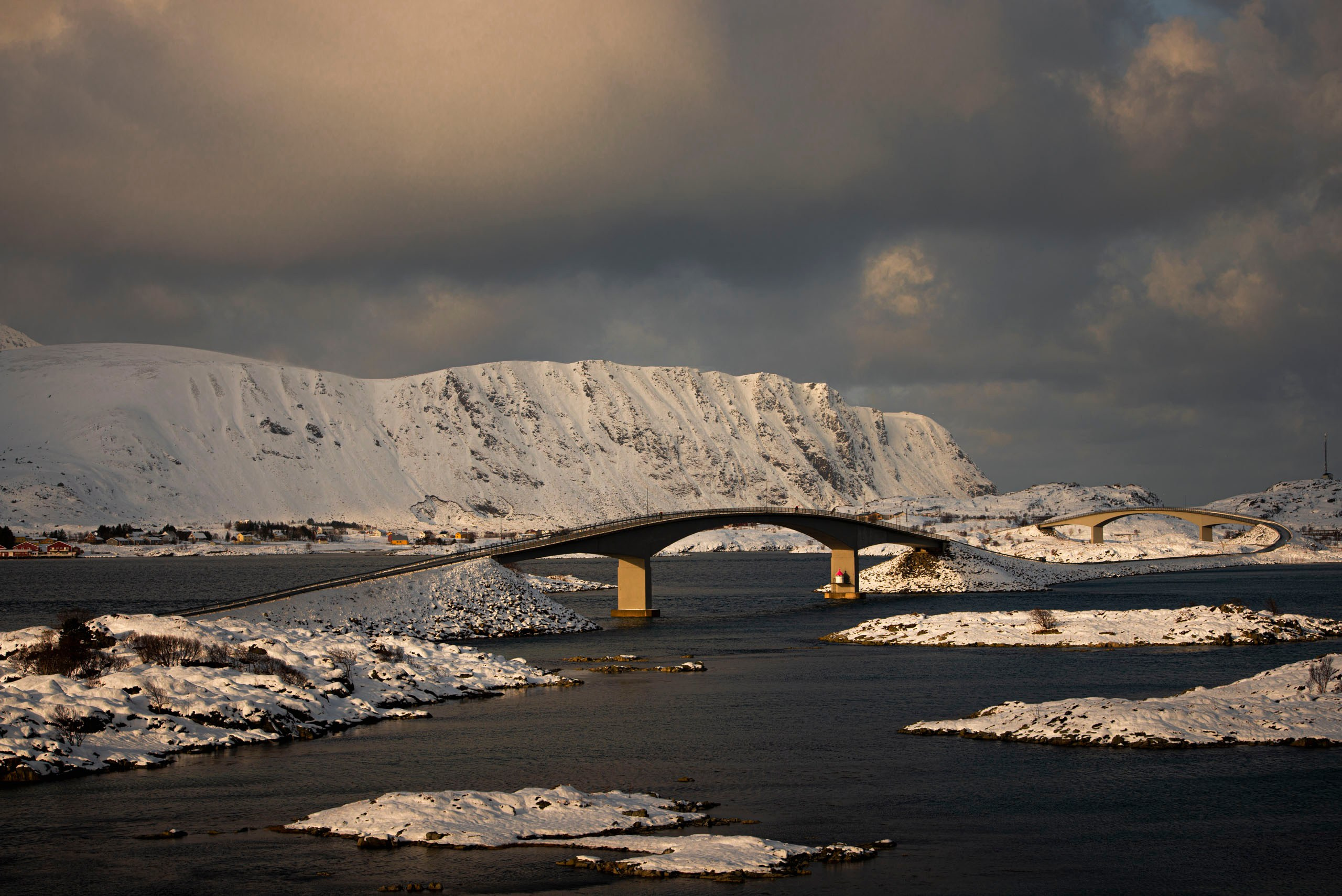 Изогнутые мосты Фредванга (Fredvang Bridges) соединяют заснеженные острова Лофотен в Норвегии на фоне гор и пасмурного неба.