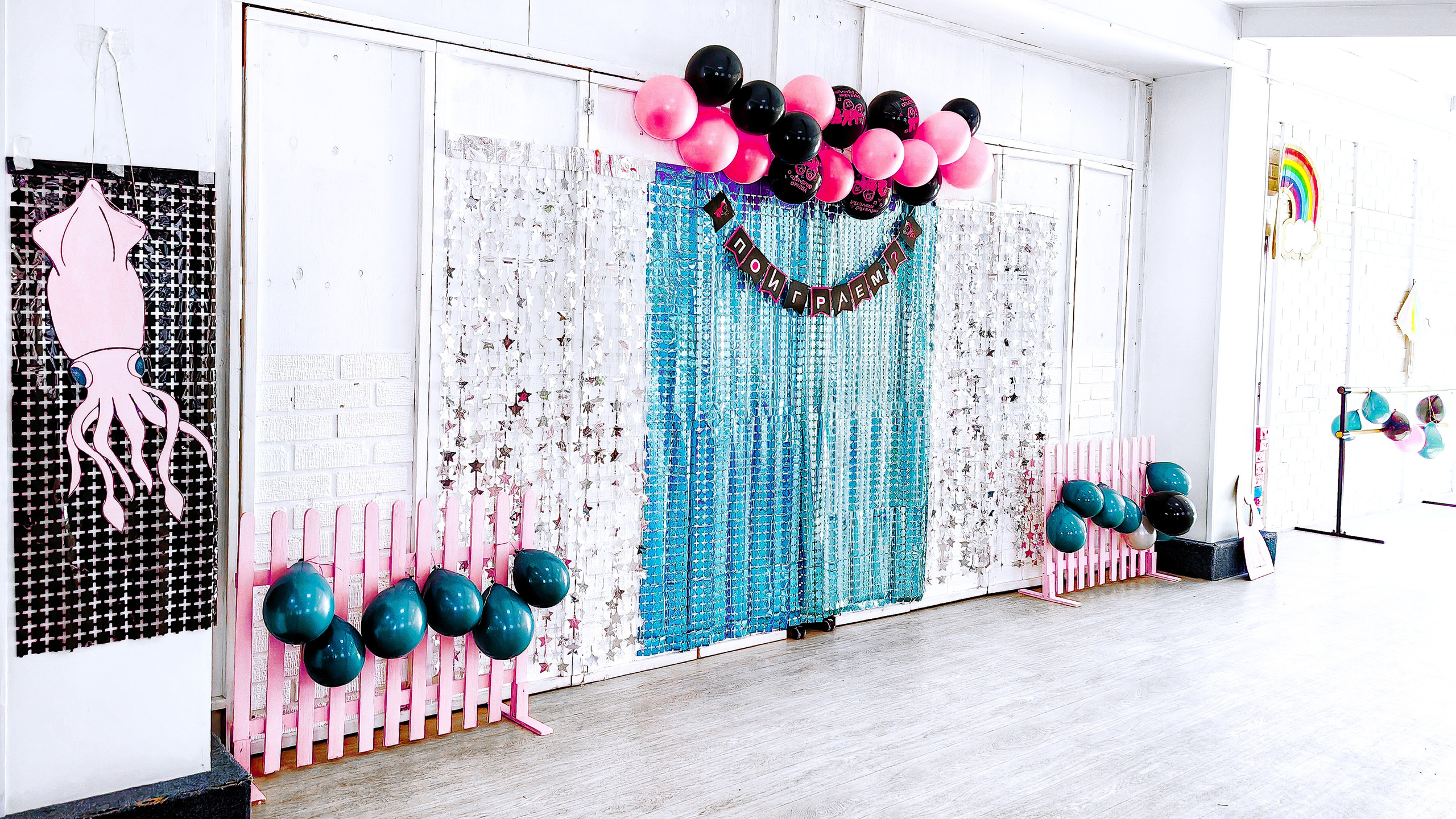 Зал оформлен для вечеринки в стиле игры в кальмара: на стенах голографический бирюзовый и черный фоны, серебряные звезды, розовые заборы, зеленые, розовые и черные шары. Вдали цветные картонные фигуры радуги и зонтика.