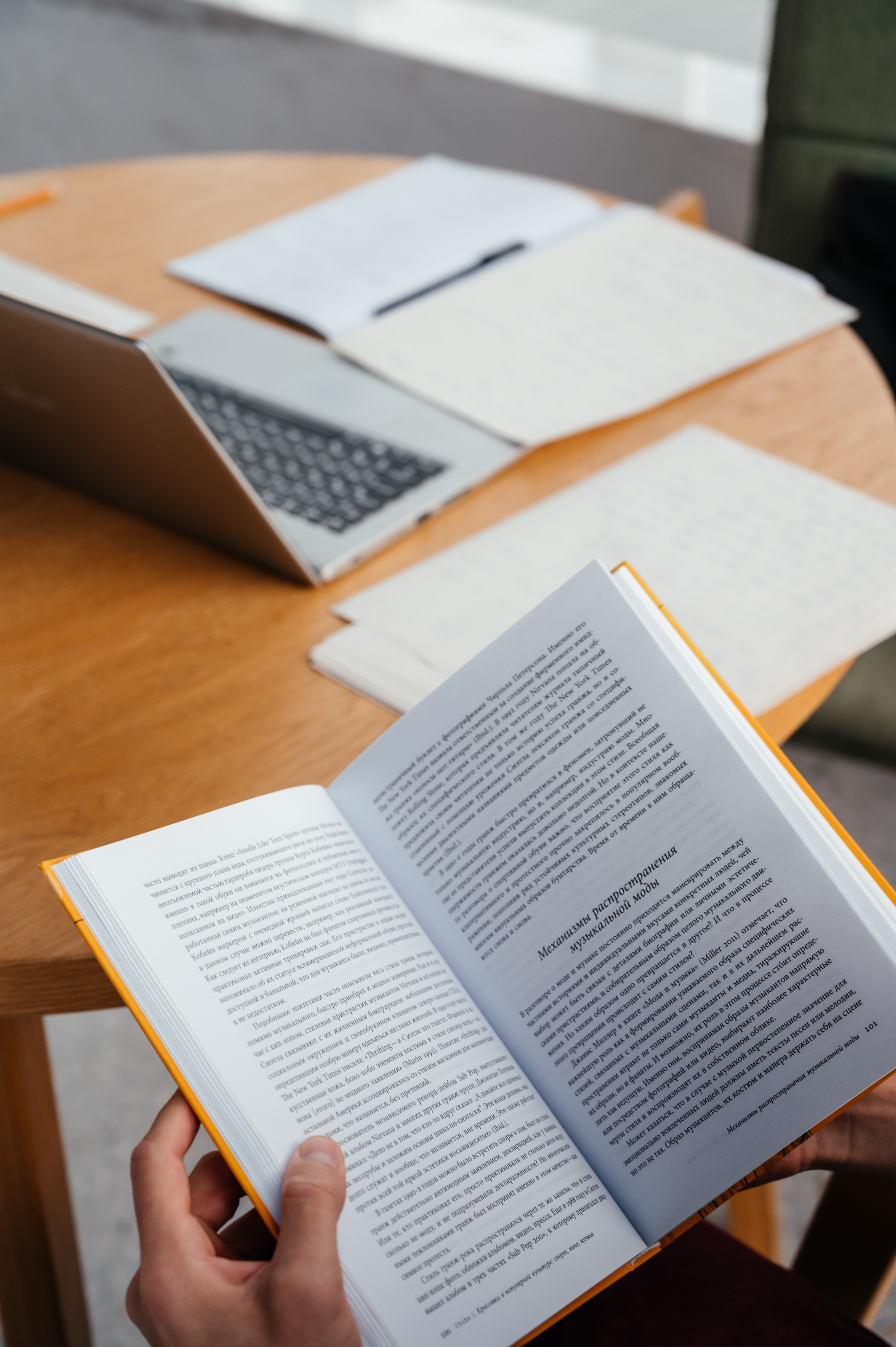 на переднем плане раскрытая книга в руках, на заднем плане на столе ноутбук и тетрадка