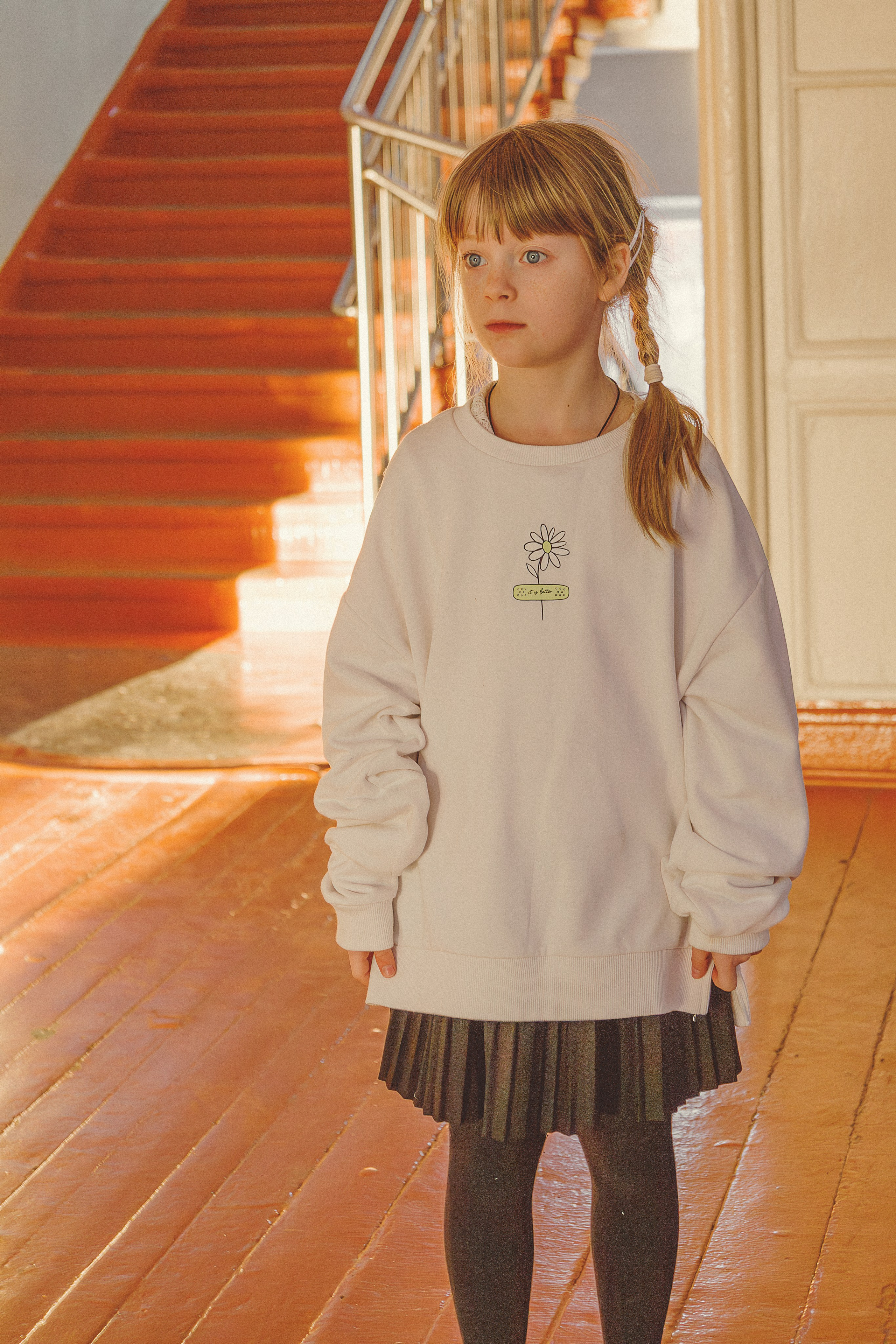 Девочка с заплетенными в косу волосами, в белой толстовке безразмерного размера и черной юбке стоит на залитой солнцем деревянной лестнице в школьном коридоре. Выражение ее лица задумчивое.