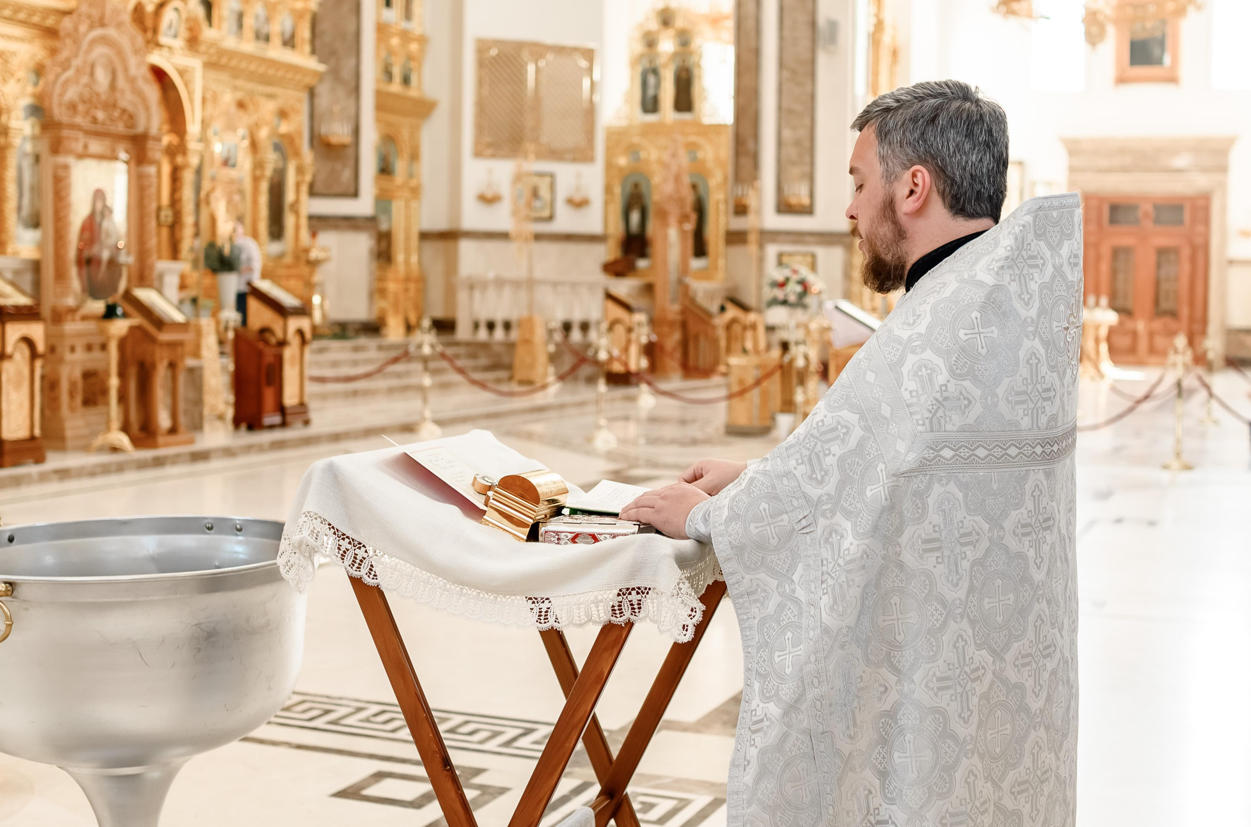 Святой отец читает молитву возле купели в храме с белыми стенами.