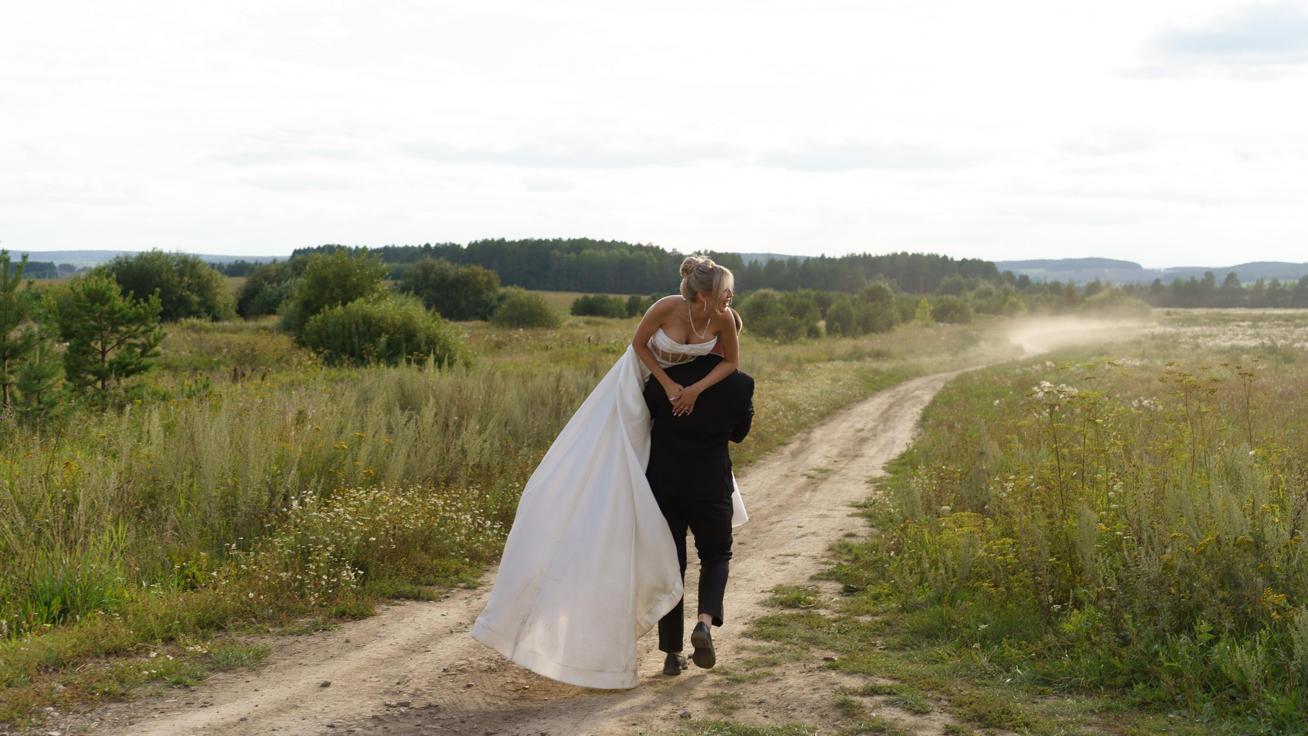 Жених с любовью несёт невесту по просёлочной дороге — её белоснежное платье развевается на ветру, а вокруг раскинулся бескрайний летний пейзаж с цветущими лугами | свадебная фотосессия на закате Екатеринбург
