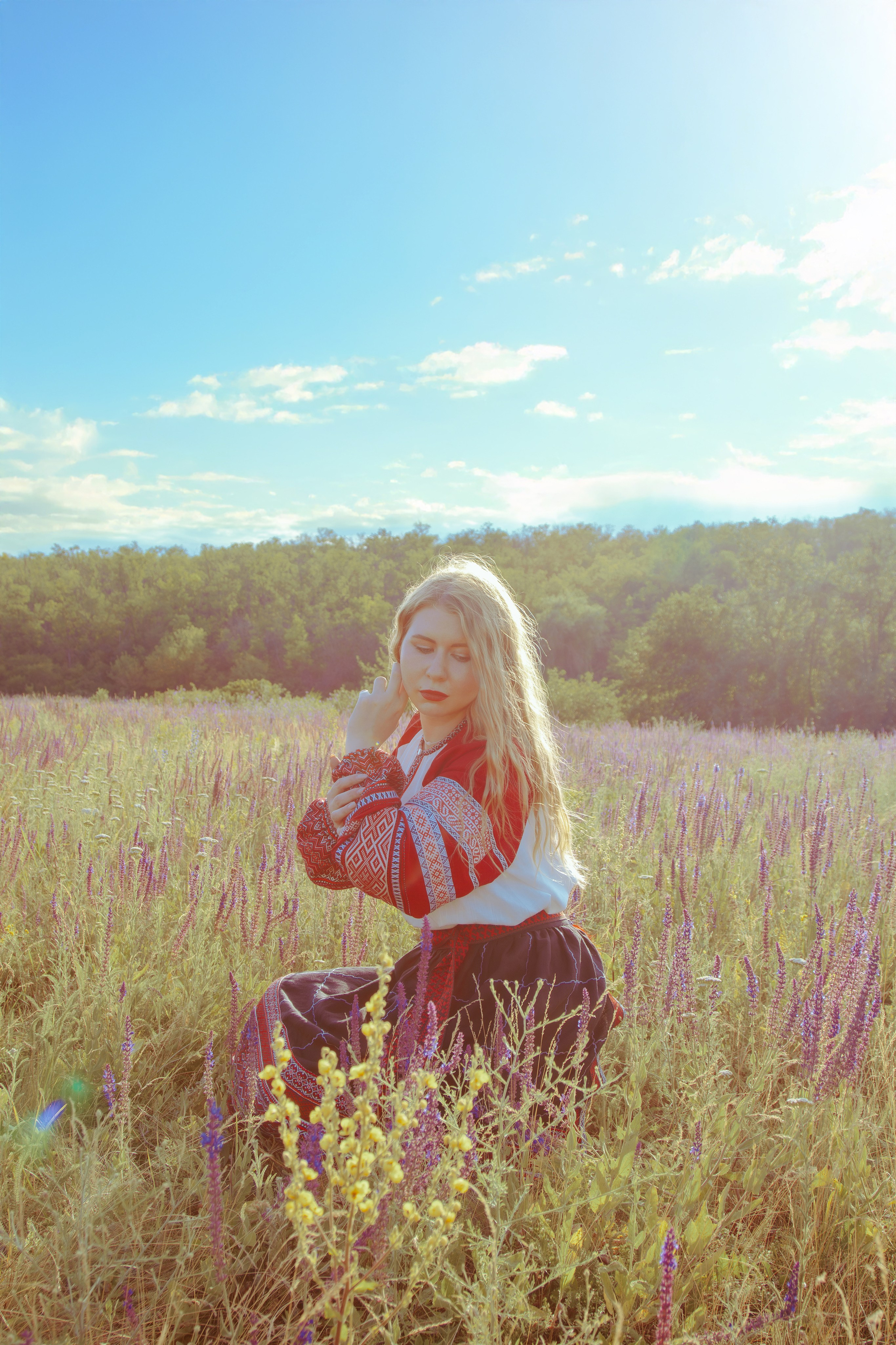 Девушка в славянском платье сидит на коленях в поле при солнечном свете