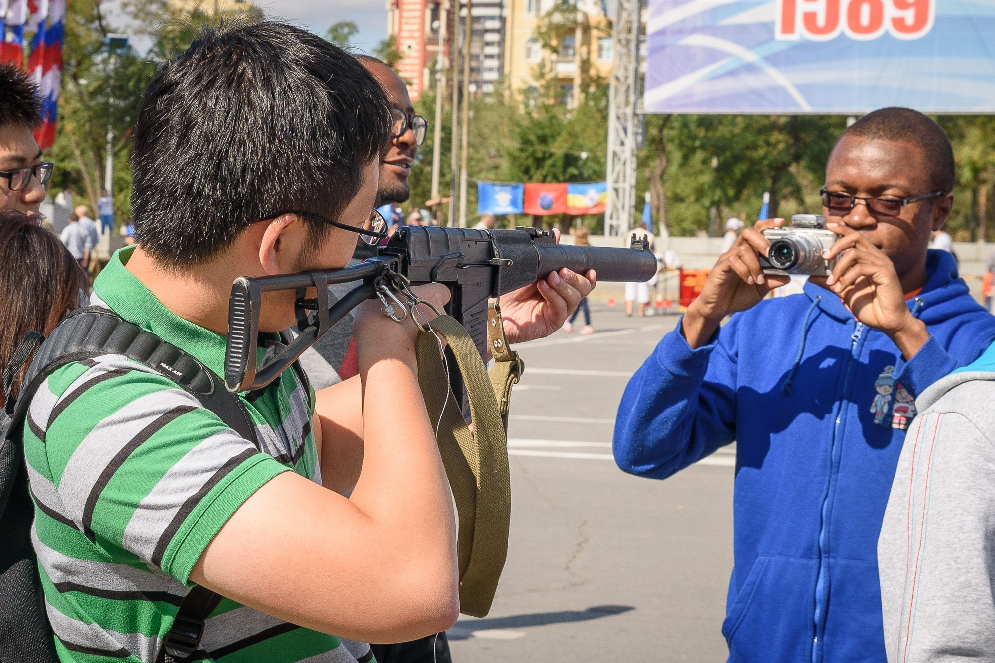 Фотограф в синей куртке снимает мужчину, который целится снайперской винтовкой
