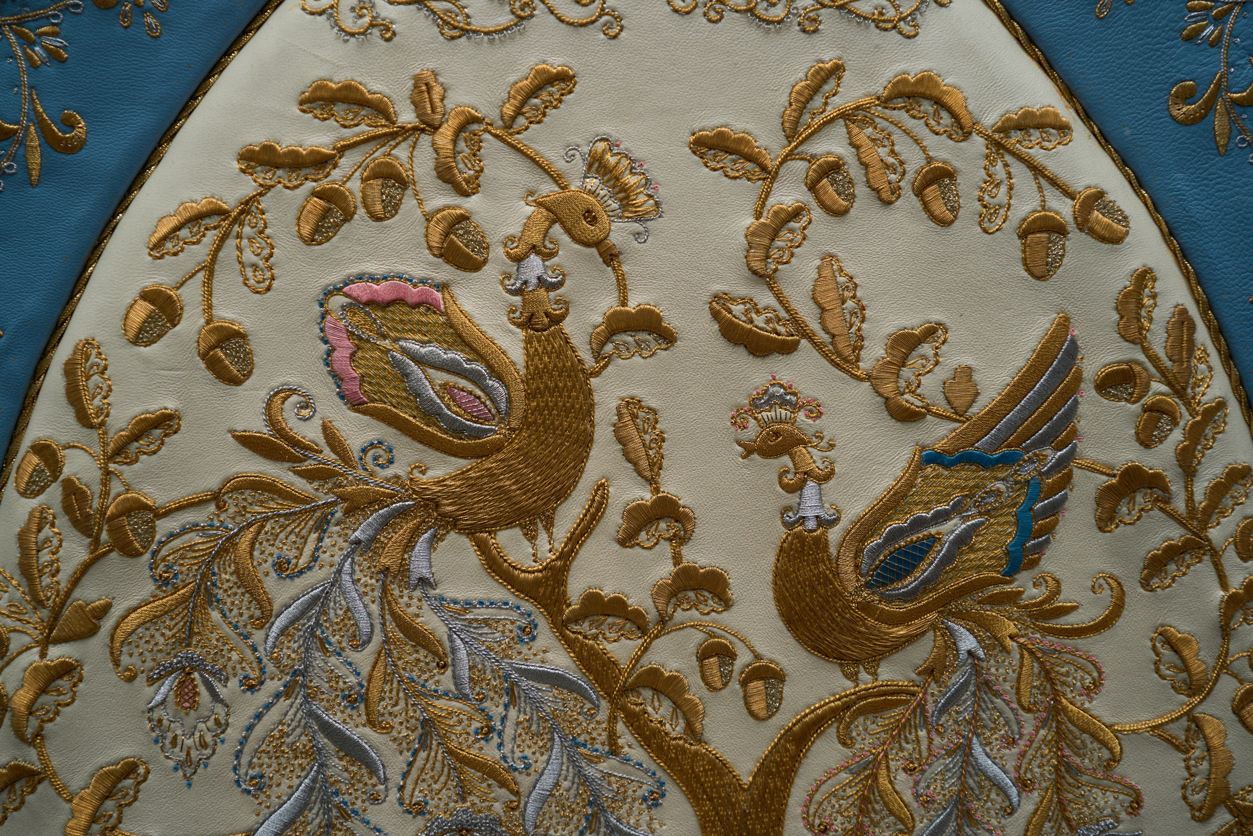 Детальный фрагмент торжокской золотошвейной вышивки, представленный в Музее торжокских золотошвей в Торжке. Вышивка выполнена на светлом фоне с использованием золотых, серебряных и цветных нитей, изображающих пышные растительные орнаменты и стилизованных птиц, включая павлина и других экзотических птиц
