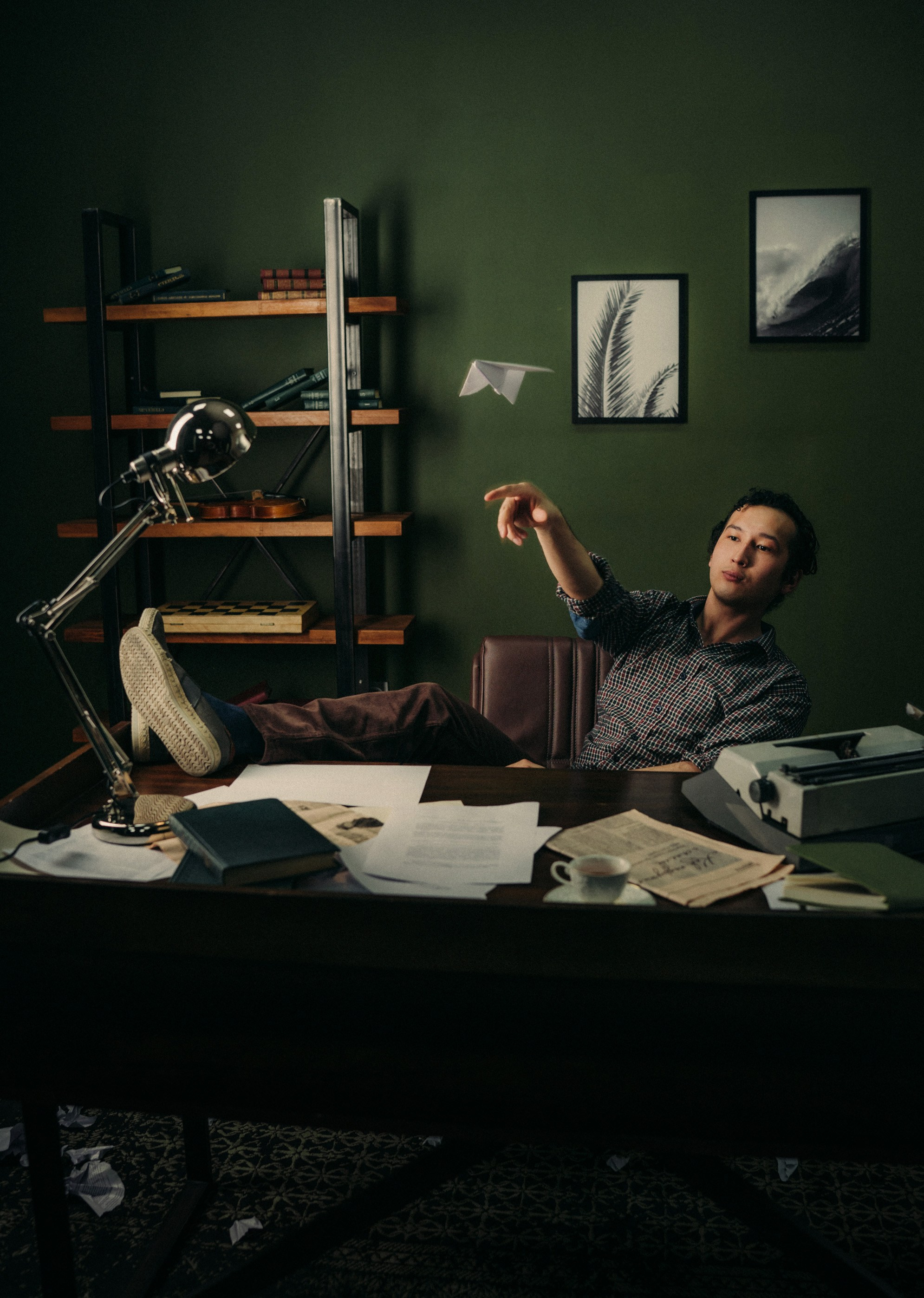 Мужчина в клетчатой рубашке сидит за письменным столом и запускает бумажный самолётик в зелёном кабинете