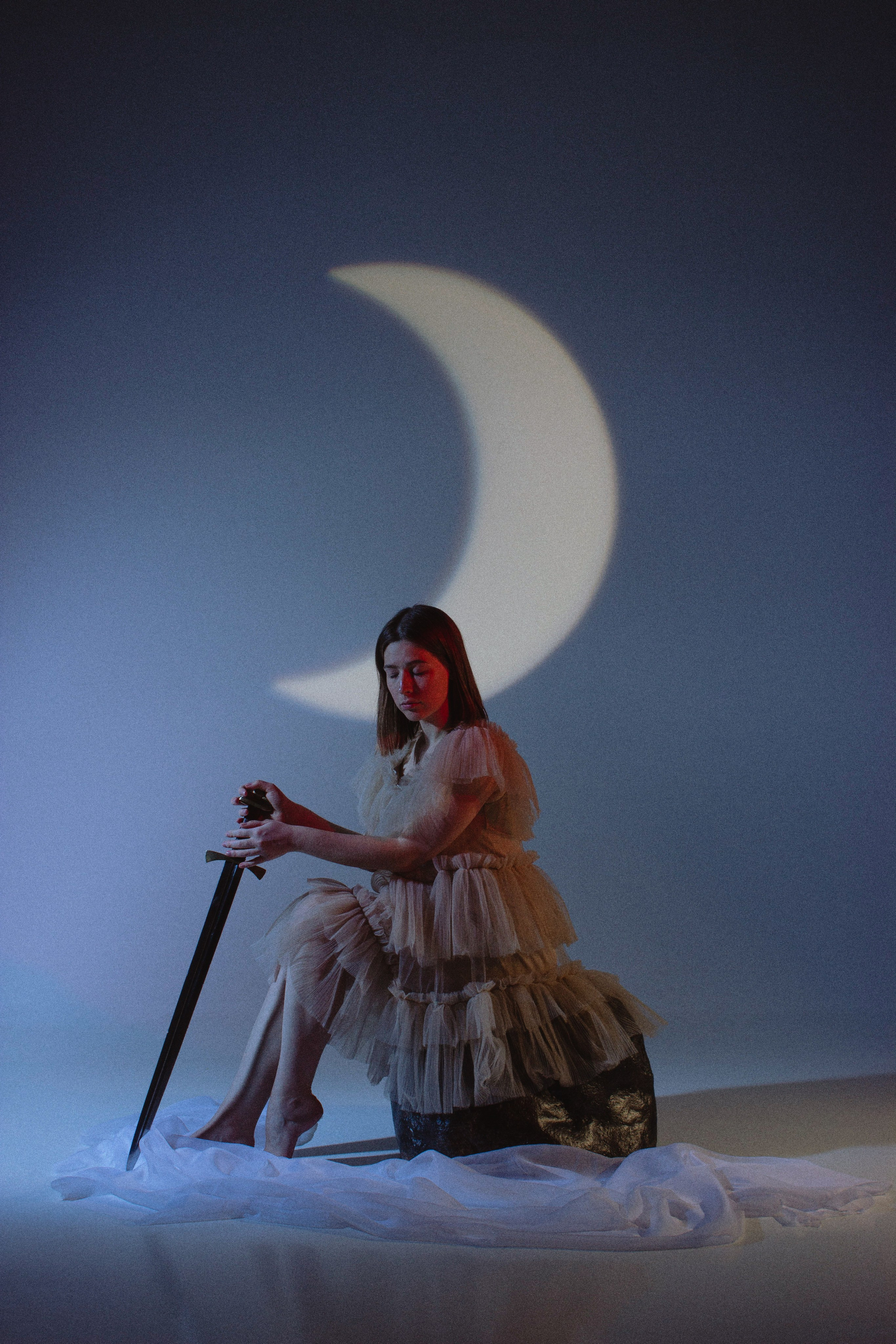 Студийный портрет девушки в платье и с мечом в руках на фоне силуэта полумесяца