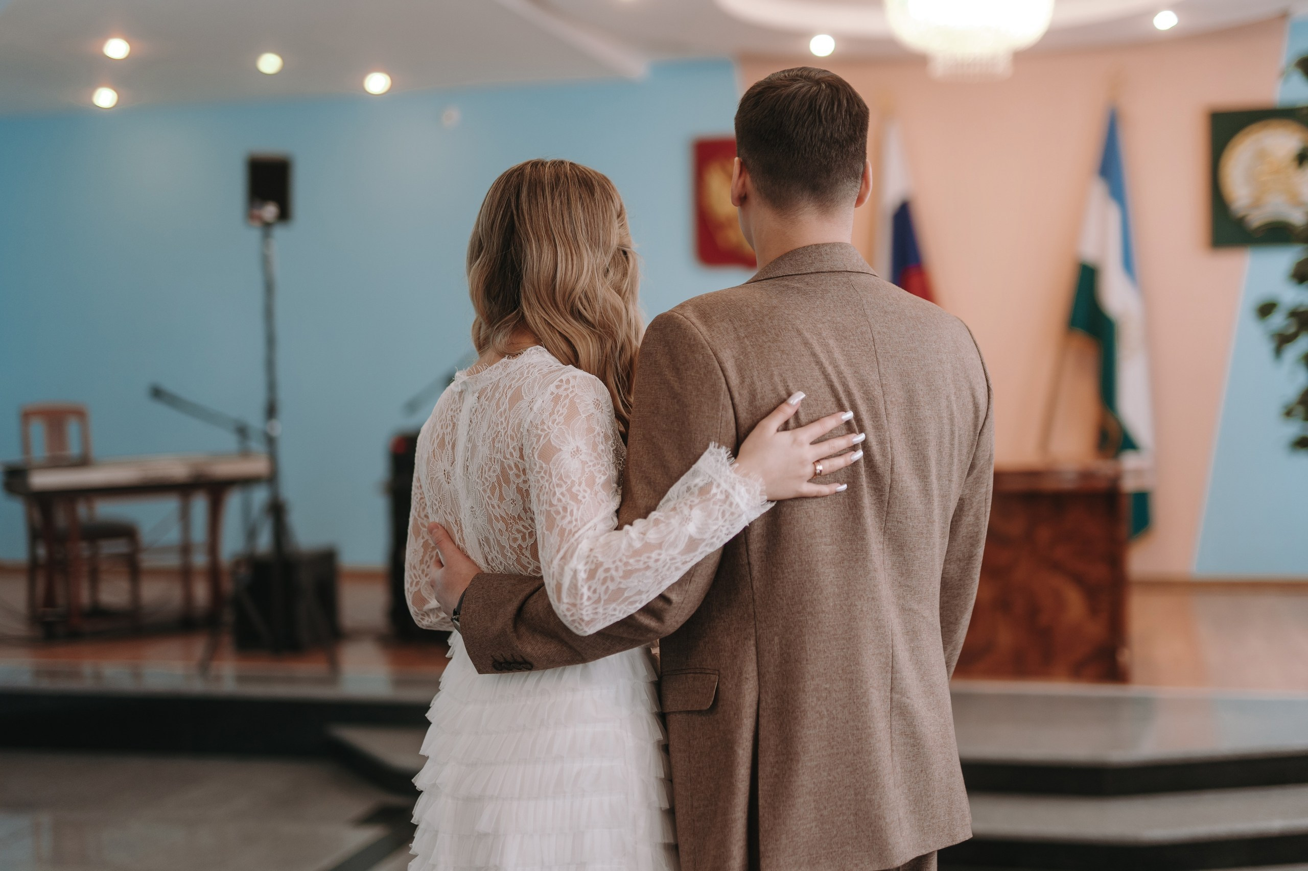 Молодожены Эмиль и Даша стоят спиной к камере, обнявшись, на регистрации брака в Октябрьском загсе.
