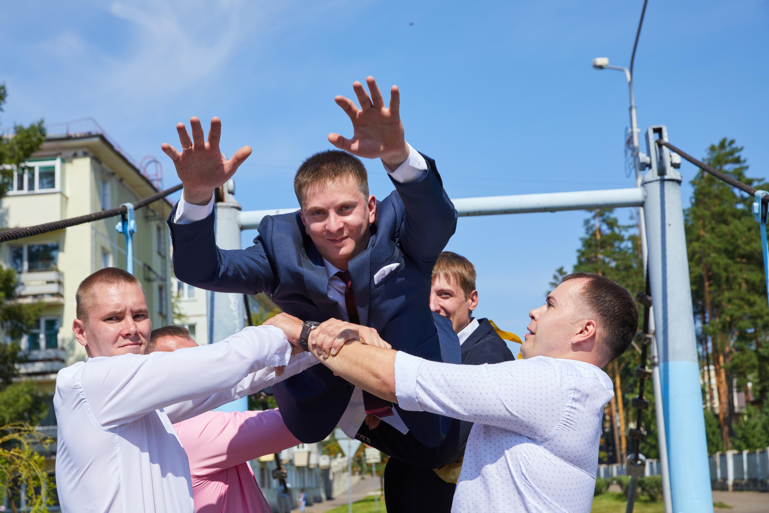 Друзья жених подкидывают его вверх, на заднем плане подвесной мост