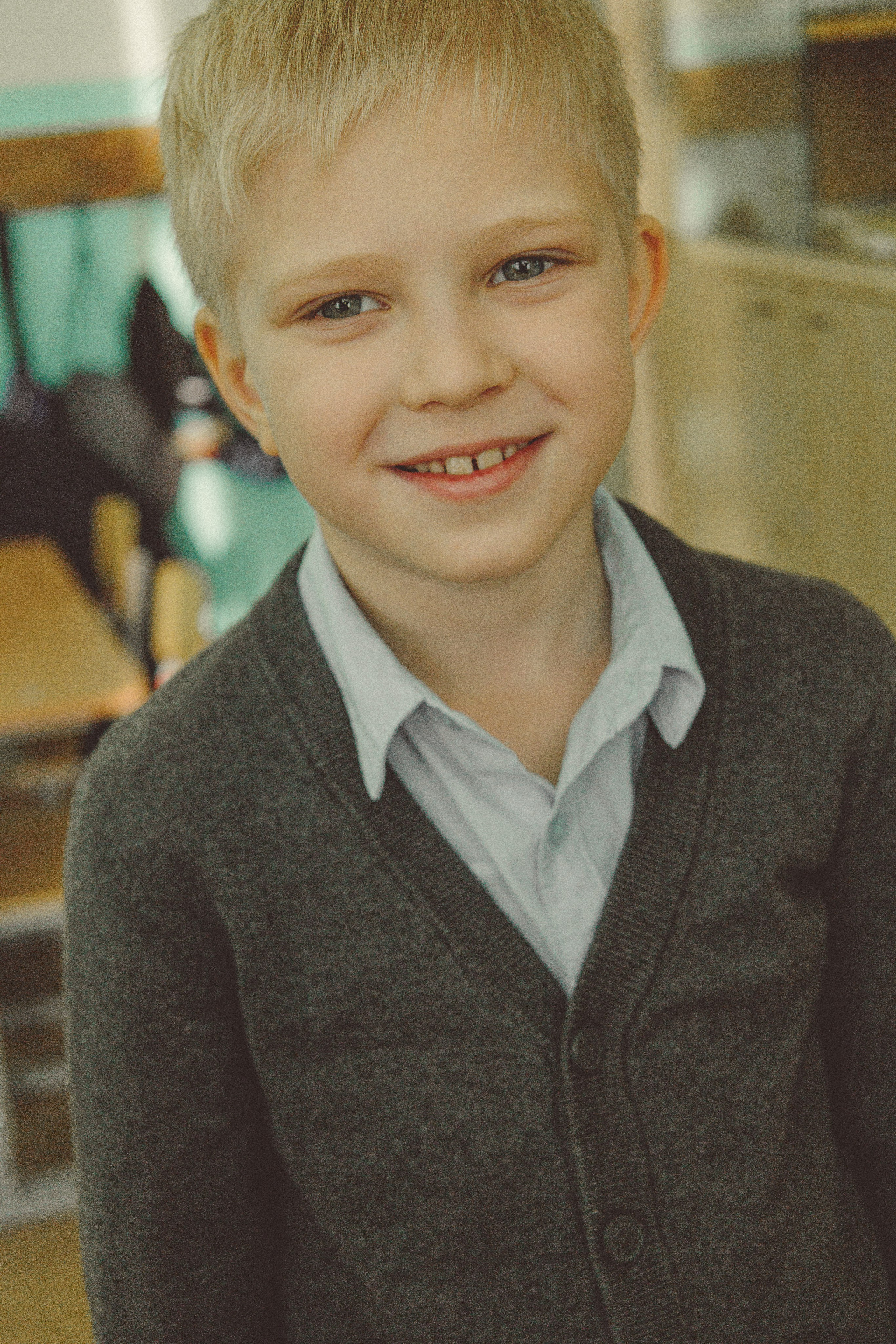 Улыбающийся светловолосый мальчик в светло-голубой рубашке и темном кардигане стоит в классе. Теплый солнечный свет создает дружелюбную, веселую атмосферу.