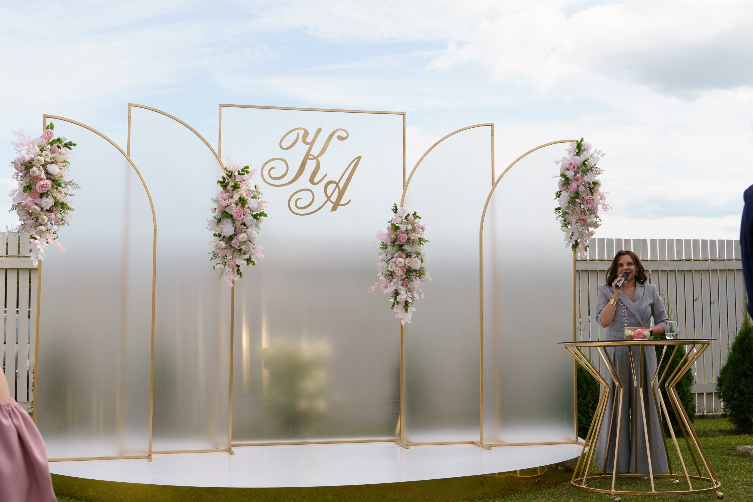 Элегантная свадебная арка с инициалами «КА», украшенная пышными композициями из нежно-розовых и белых цветов, на фоне светлого неба — изысканный фон для торжественного момента | репортажная свадебная фотосъемка Екатеринбург