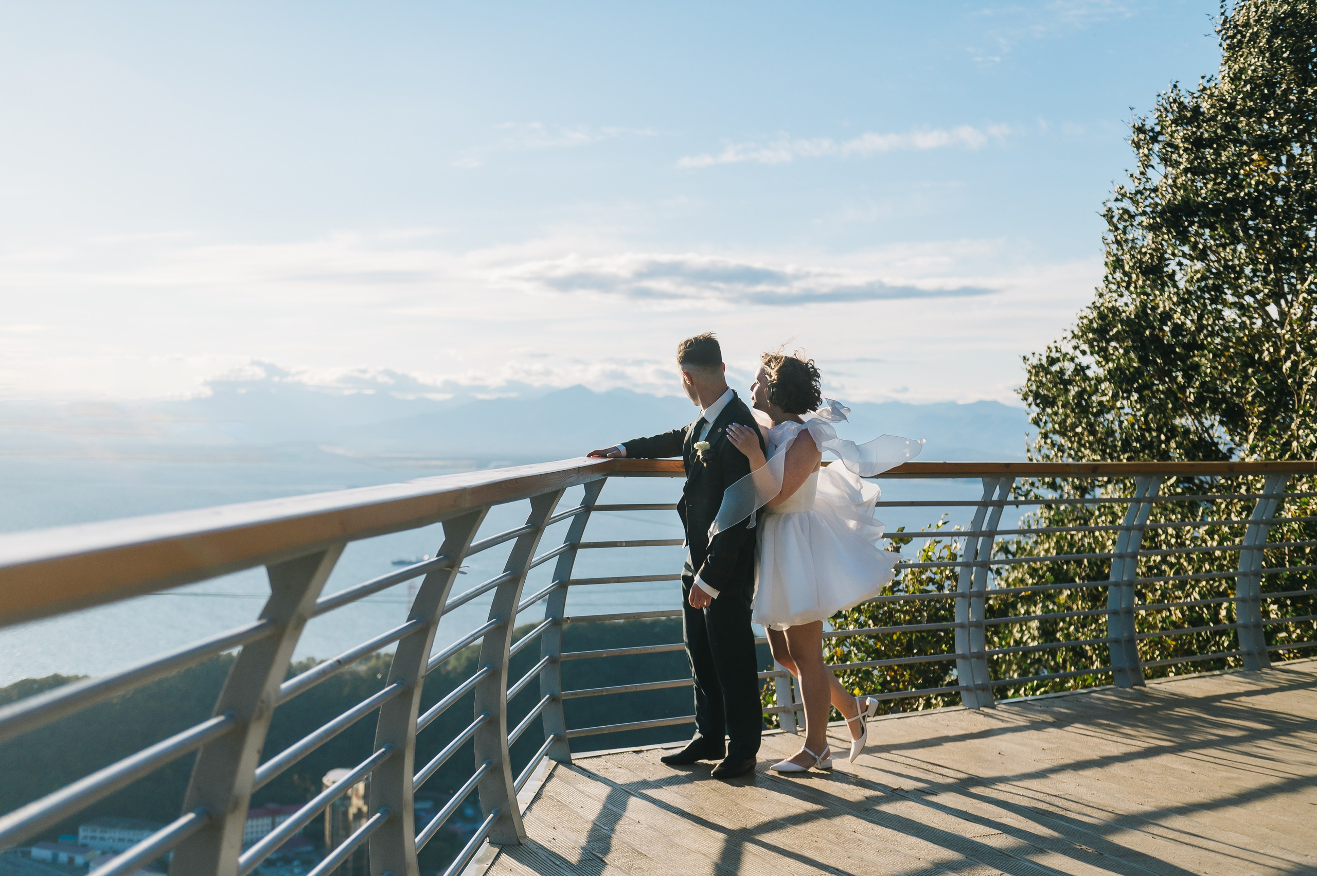 Жених в костюме и невеста в белом платье стоят на смотровой площадке с видом на горы и воду. Атмосфера романтичная, светлая.