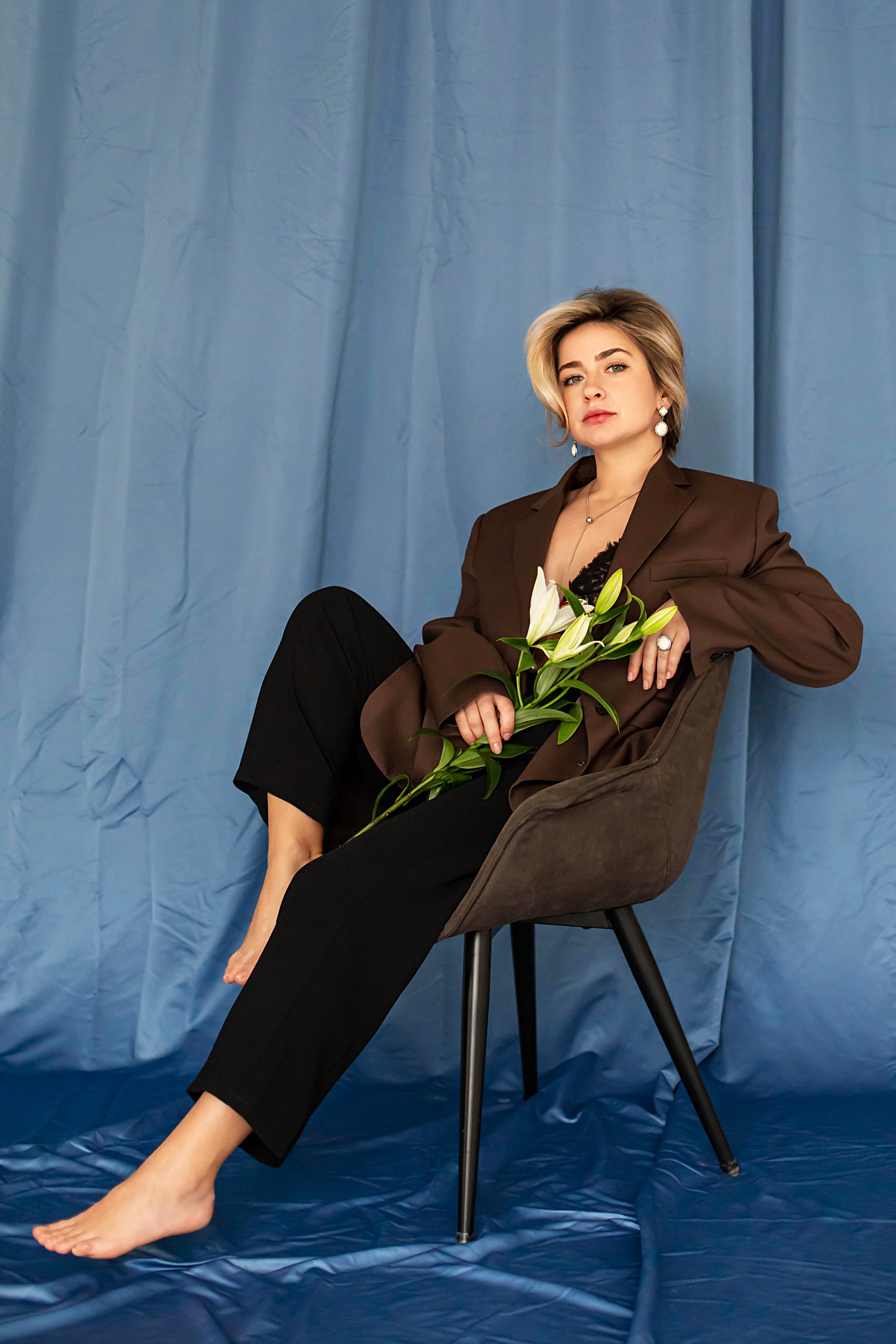 Съемка милированной блондинки на стуле в коричневом пиджаке на синем фоне с лилией в руках