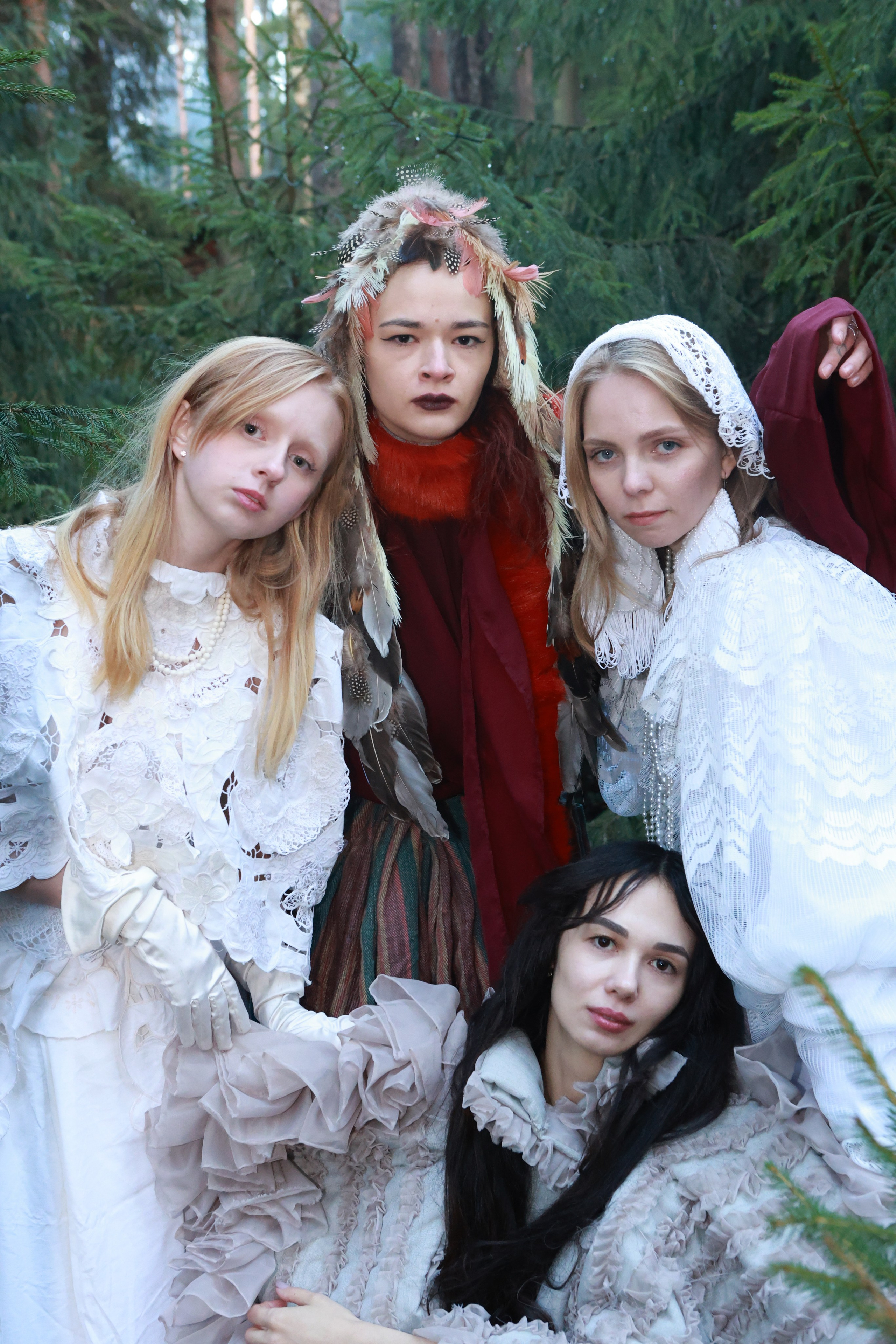 Группа из пяти моделей в винтажных нарядах с рюшами и перьями позирует в хвойном лесу. Соединённые руки, экстравагантные образы, атмосферная арт-съемка. Профессиональная съемка для модных проектов.