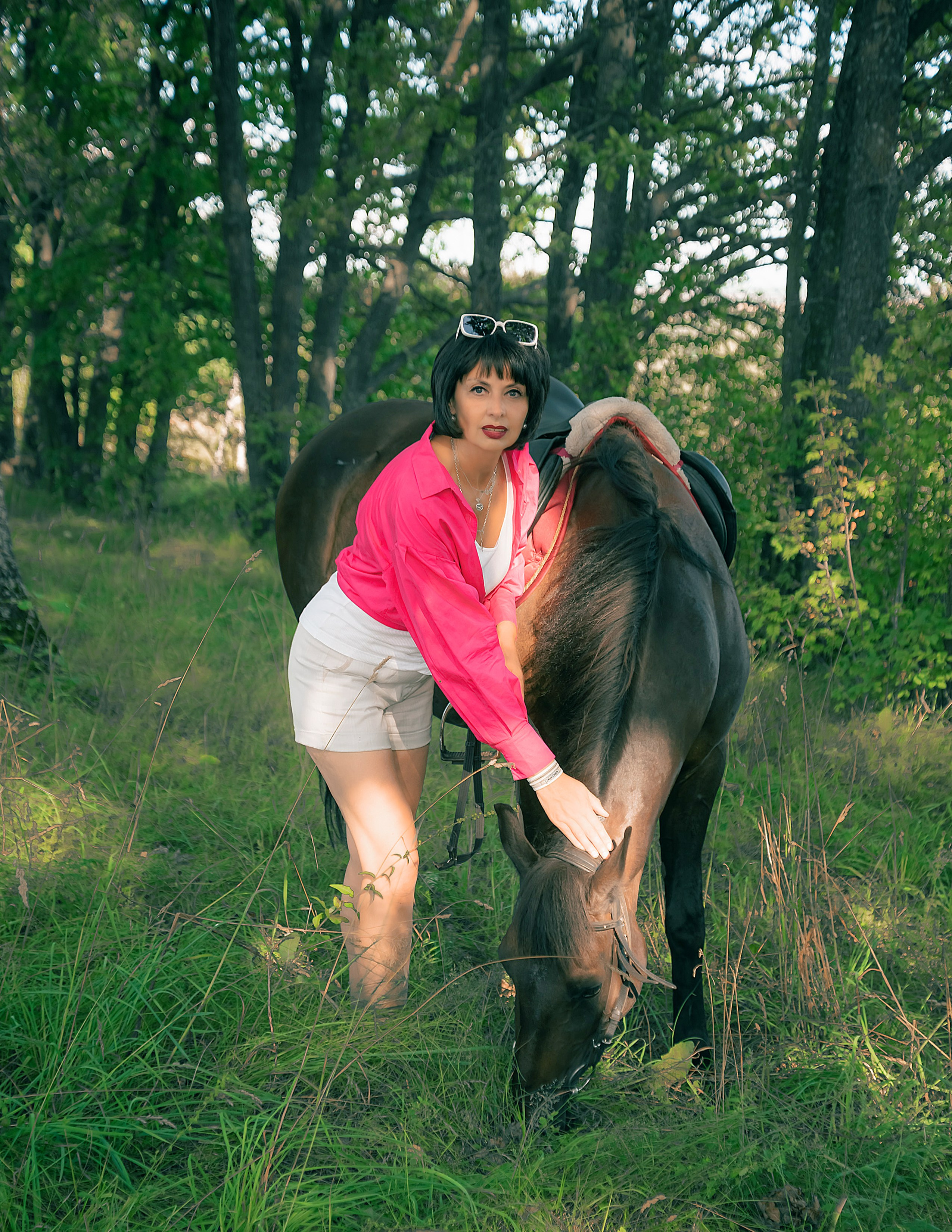 На фото женщина в рубашке и белых шортах гладит лошадь на опушке леса.