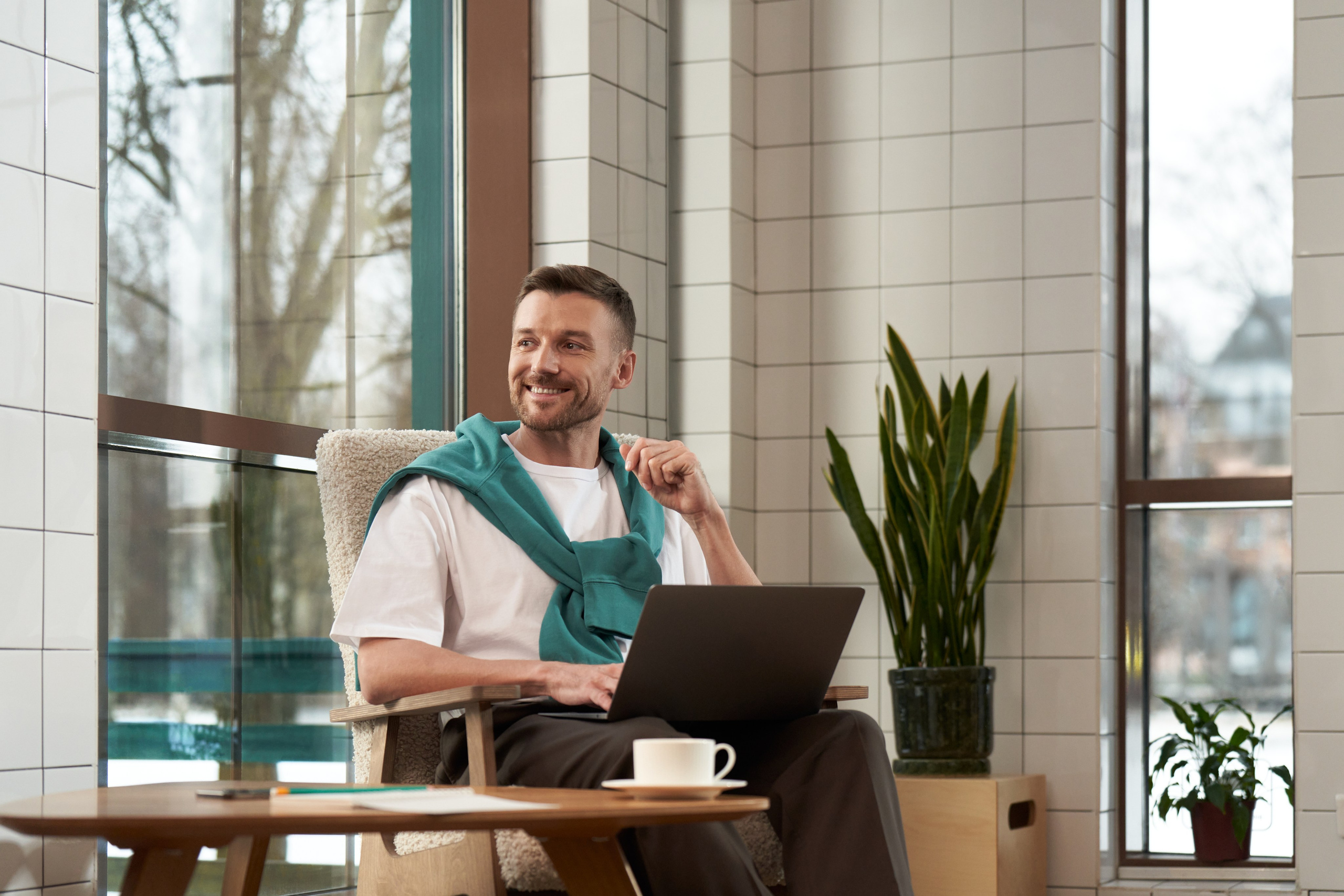 Мужчина с ноутбуком сидит в офисной лаунж-зоне у окна, расслабленная атмосфера