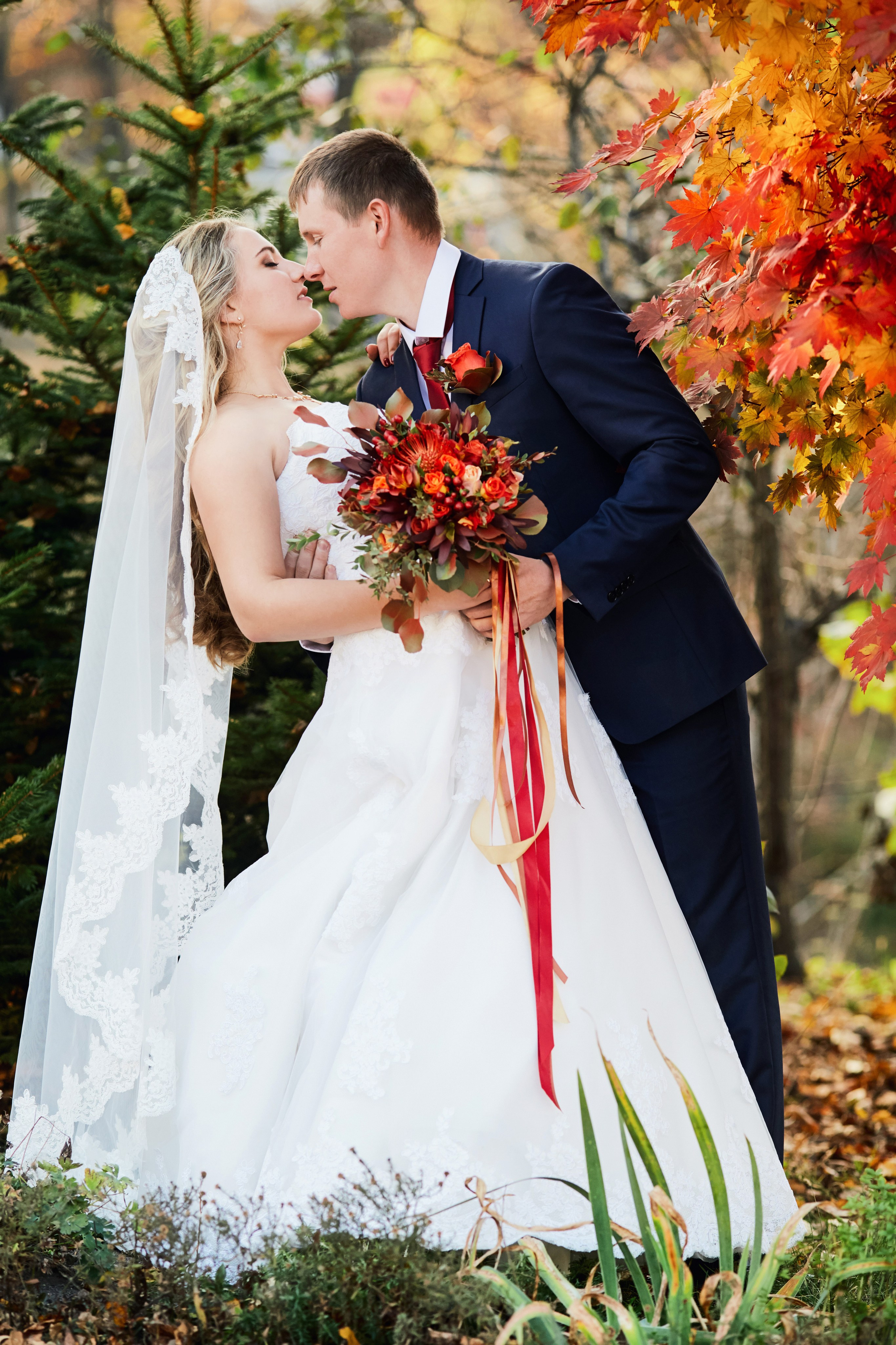 Свадебная фотосъёмка с невестой и красным букетом и женихом в классическом костюме на фоне клёна в Находке