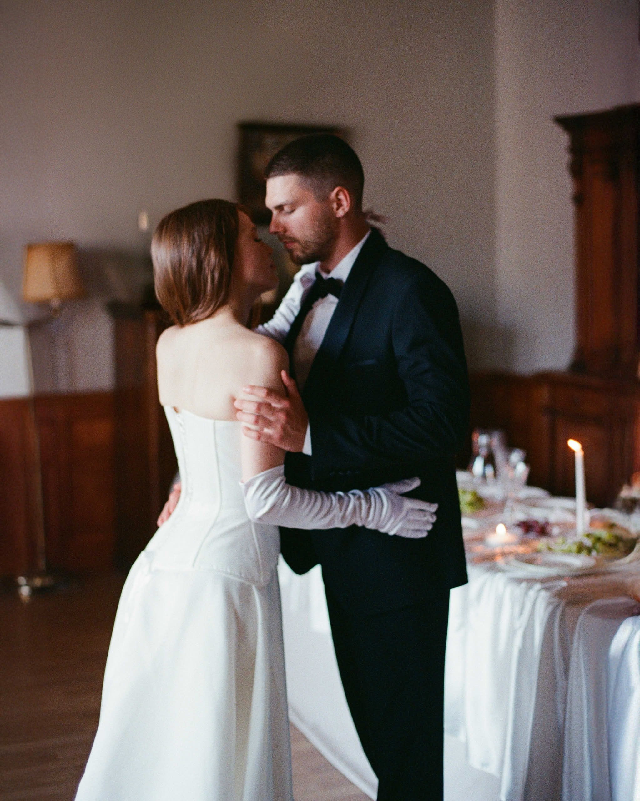 Плёночный кадр: жених и невеста танцуют у праздничного стола в тёплой камерной обстановке