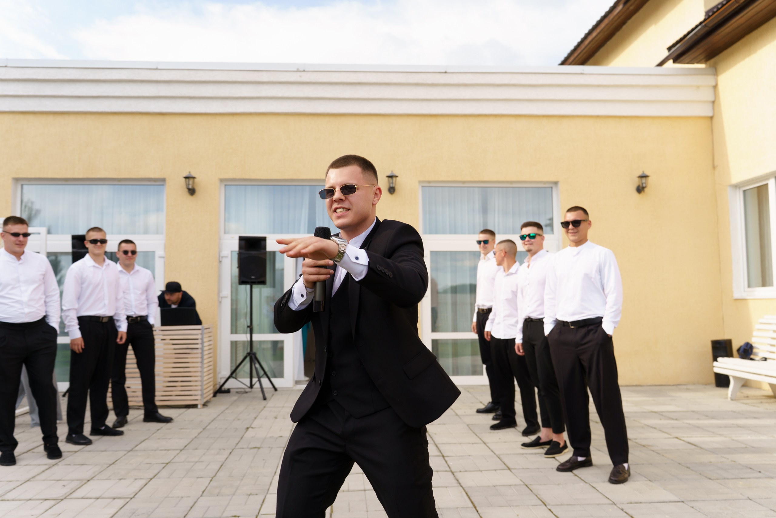 Момент искромётного выступления: жених в смокинге с микрофоном словно дирижирует свадебным торжеством, а его команда в строгих белых рубашках поддерживает динамику события на фоне уютного здания | свадебная фотосессия Екатеринбург