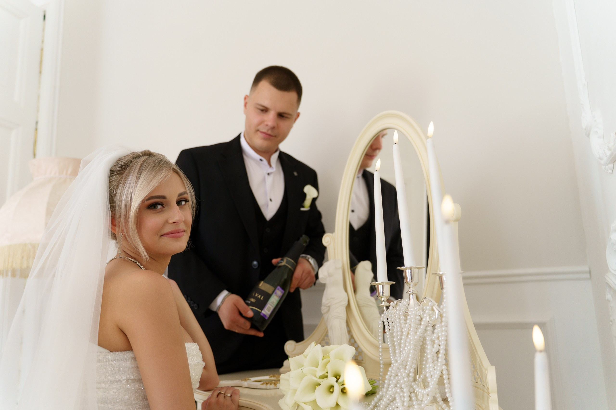 Невеста и жених в день свадьбы: невеста смотрит в камеру с лёгкой улыбкой, жених занят бутылкой шампанского. На столике — зажжённые свечи, зеркало в изящной раме и букет белых калл | свадебная фотосессия Екатеринбург