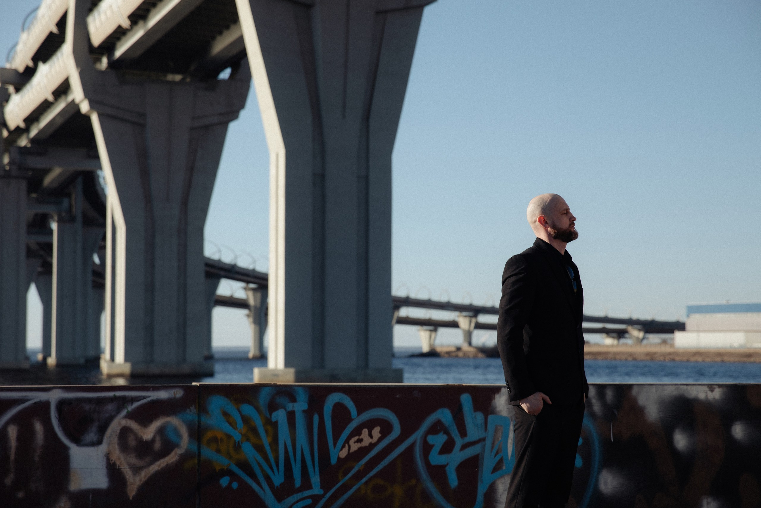 Мужчина в черном костюме у граффити и воды на фоне городской архитектуры, фотосессия в Санкт-Петербурге.