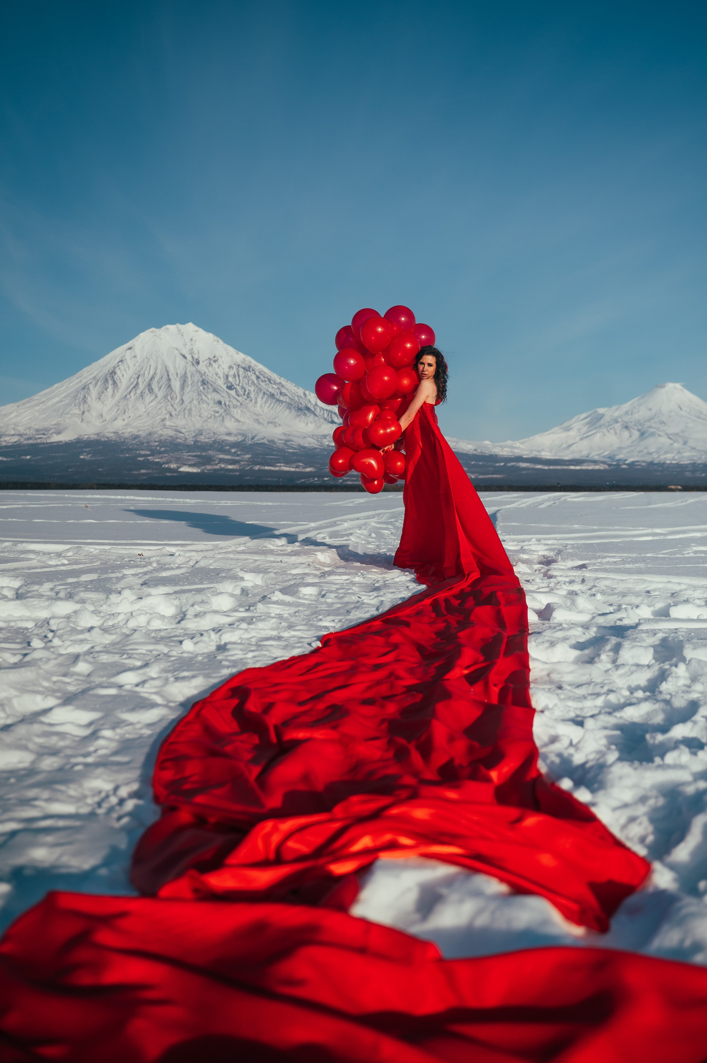 Девушка в длинном красном платье стоит на снегу с пучком красных шаров. На фоне — заснеженные вулканы, атмосфера сказочная.