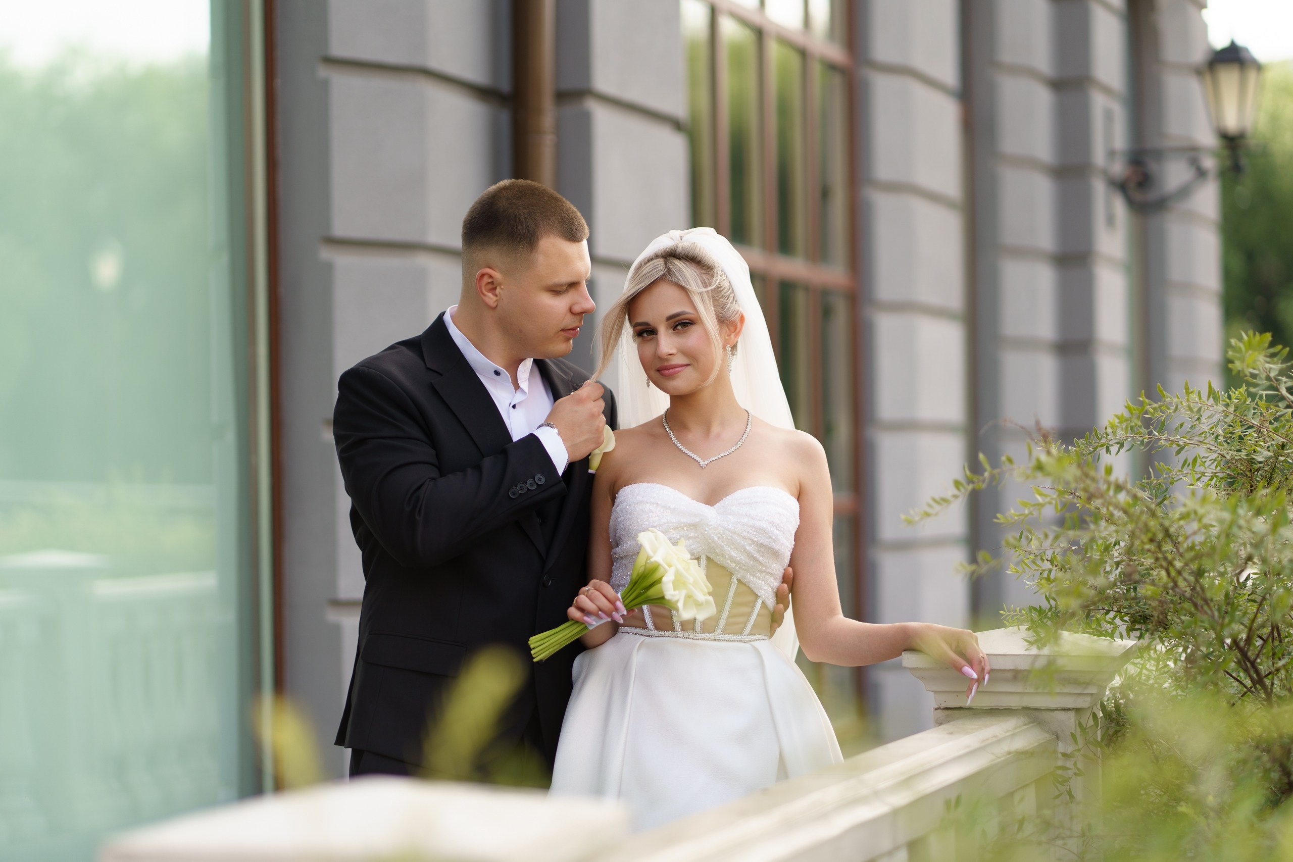 Невеста в изысканном белом свадебном платье с корсетным верхом и фатой позирует с букетом белых цветов. Рядом с ней — жених в элегантном чёрном костюме, который нежно касается её фаты. На заднем плане — здание с большими окнами и зелёными насаждениями | свадебная фотосъемка в исторических местах Екатеринбург
