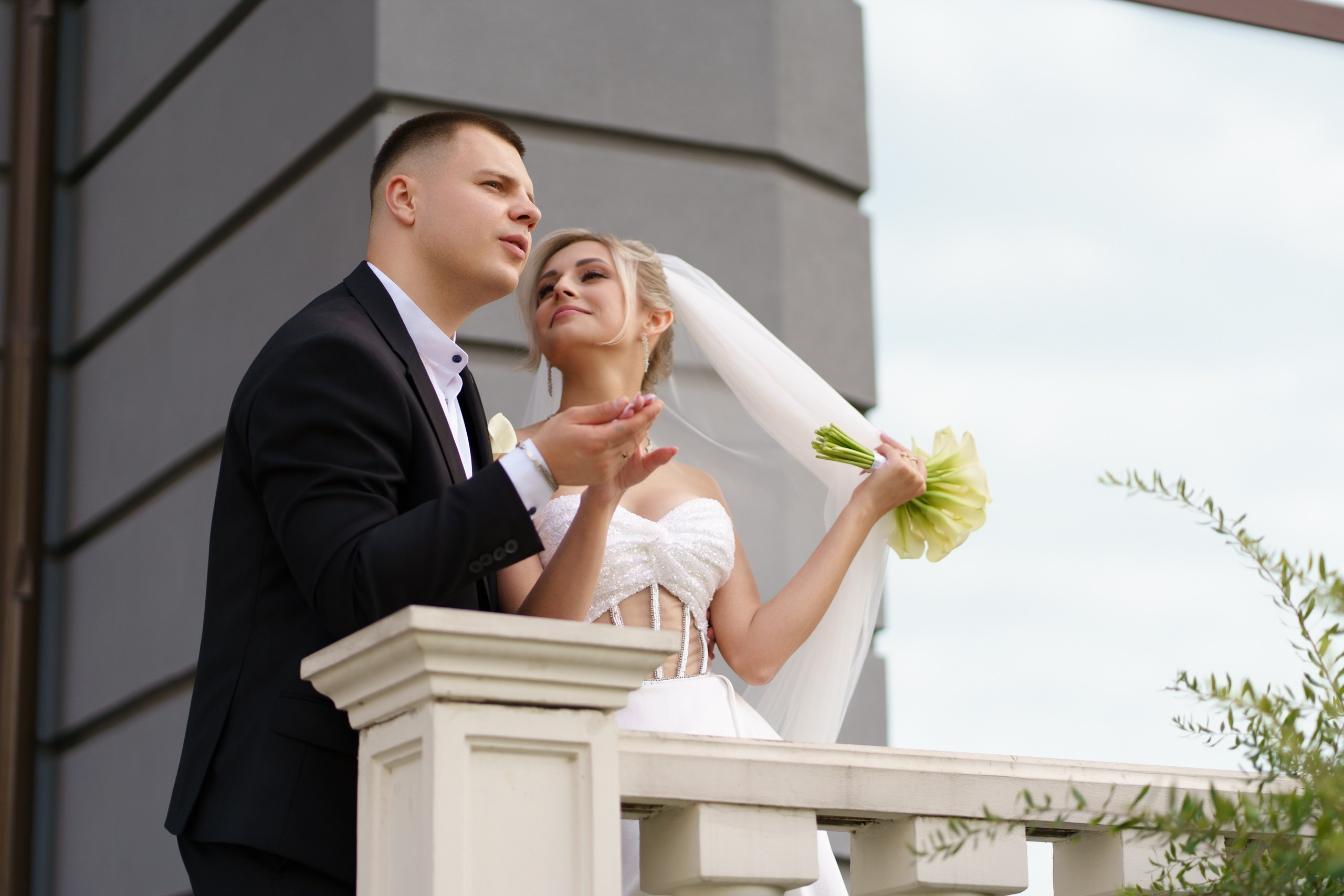 Свадебная фотосессия на балконе: жених в строгом чёрном костюме и невеста в изысканном белом платье. Невеста держит букет и слегка наклоняет голову | красивые свадебные кадры Екатеринбург