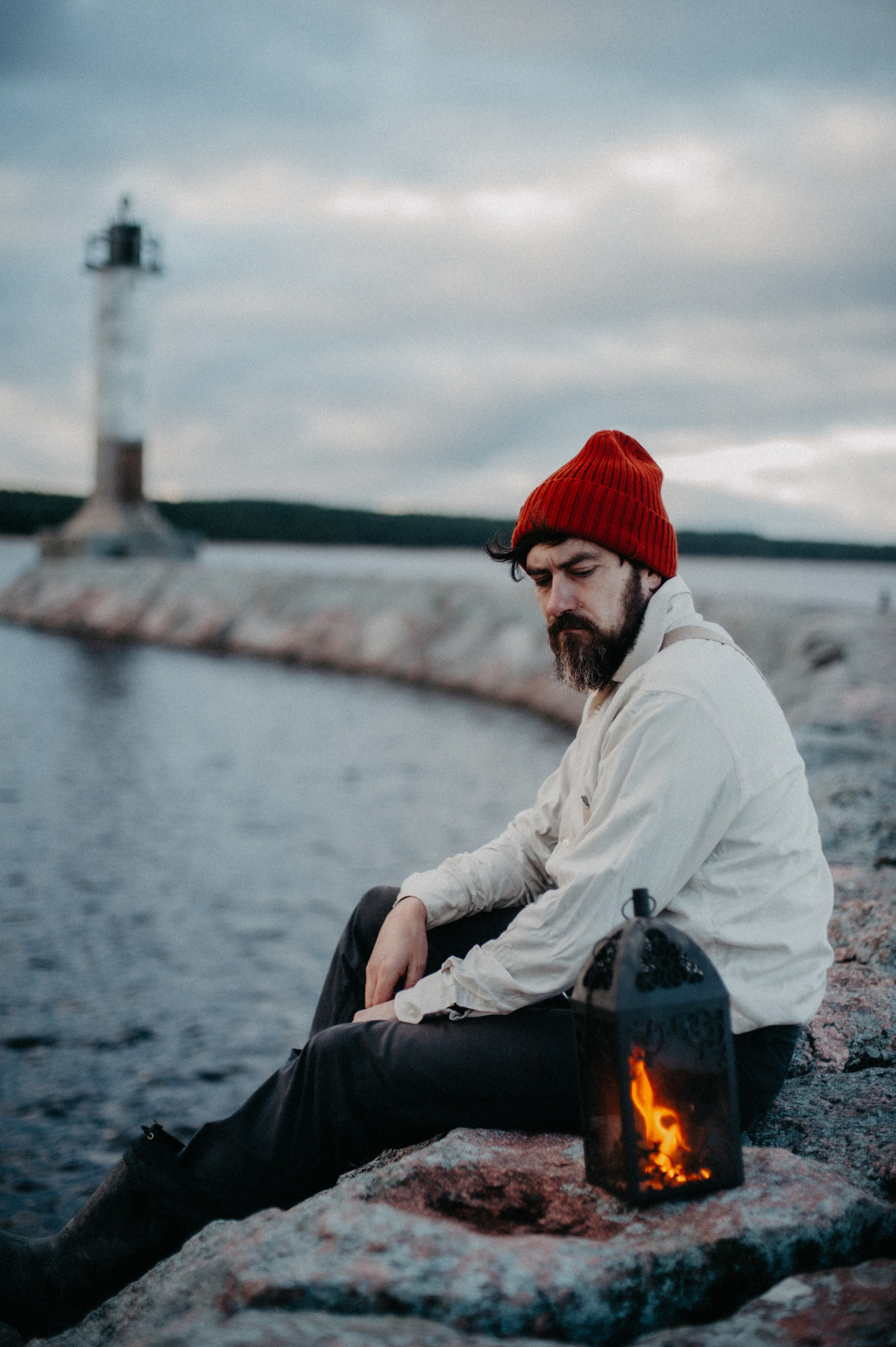 Мужчина с бородой с проседью в красной шапке сидит с фонарём у воды на фоне маяка