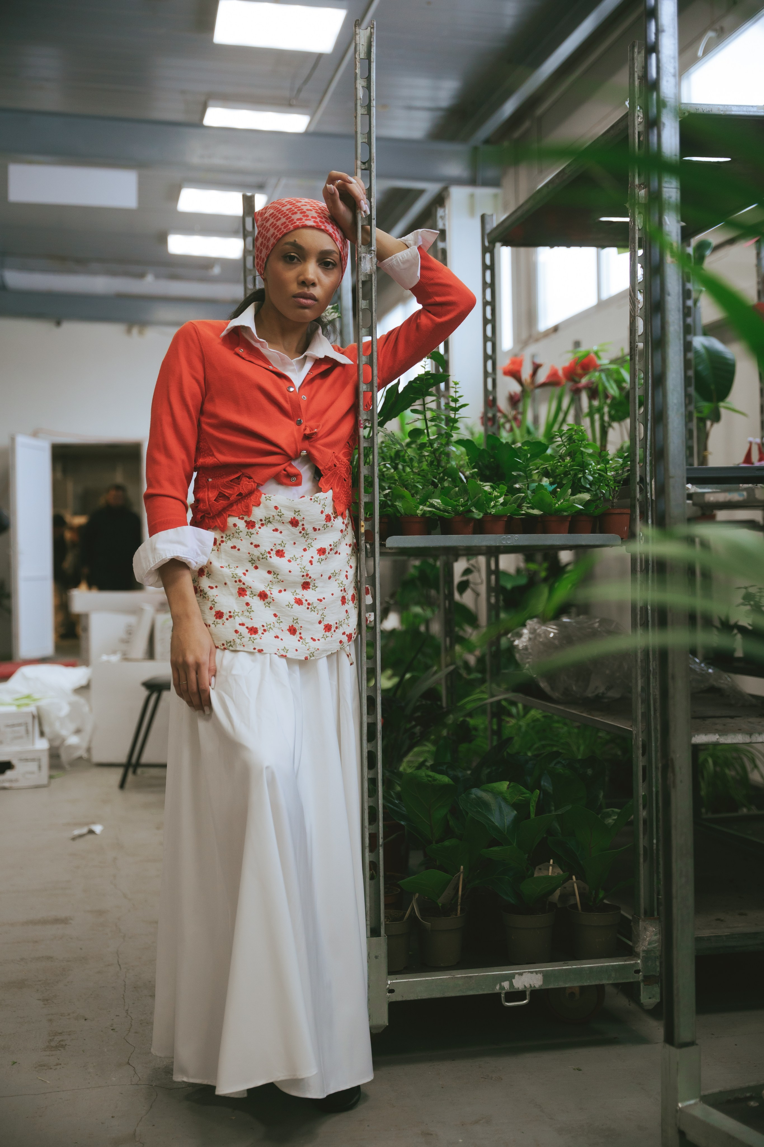 Девушка в красном кардигане и платке позирует среди стеллажей с цветами