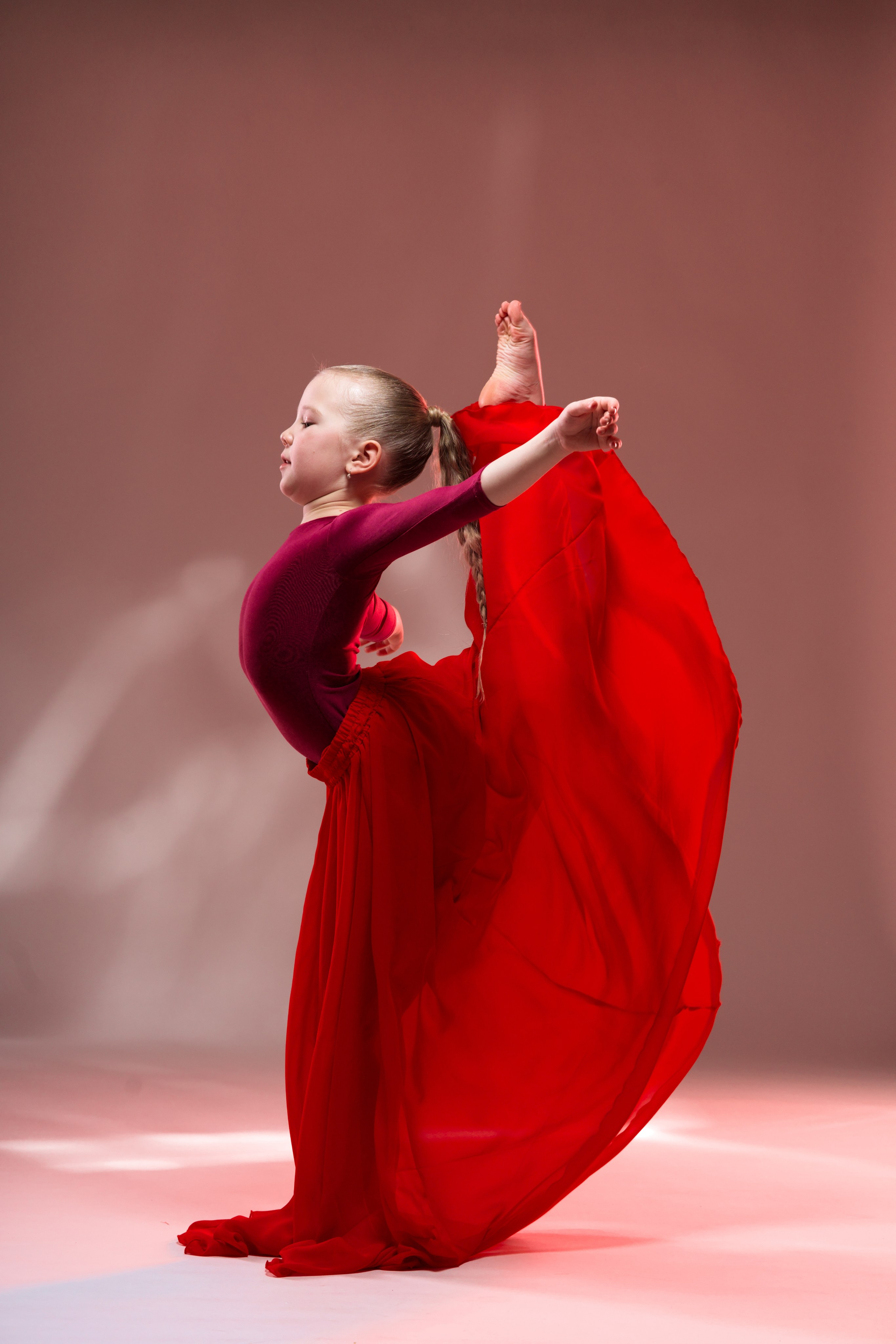 Спортивный арт-портрет гимнастки в профиль: яркое красное платье и сложная гимнастическая фигура на студийном фоне.