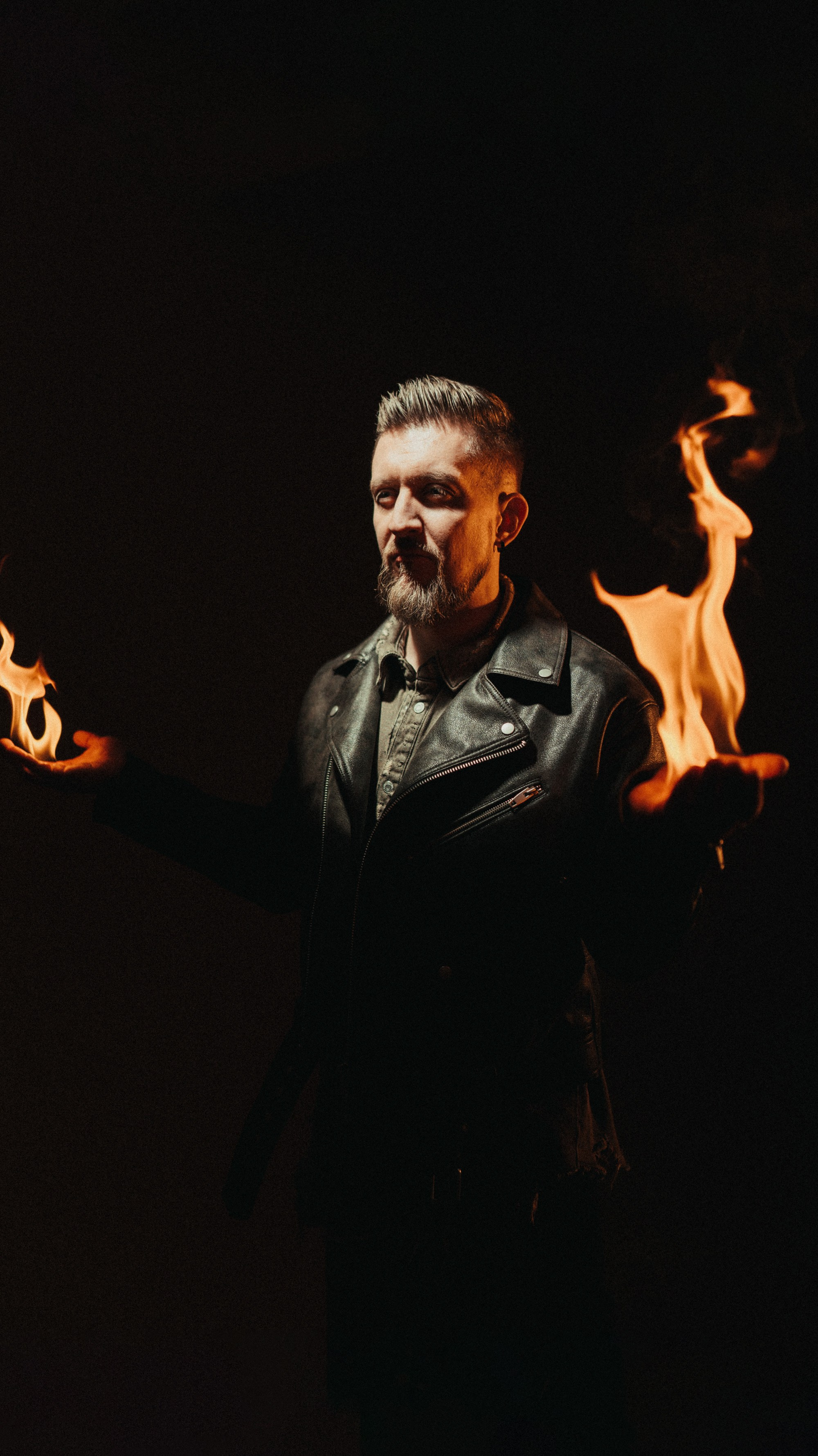 Мужчина с короткой стрижкой и бородой в чёрной кожаной куртке стоит в тёмной комнате, держа в руках огонь