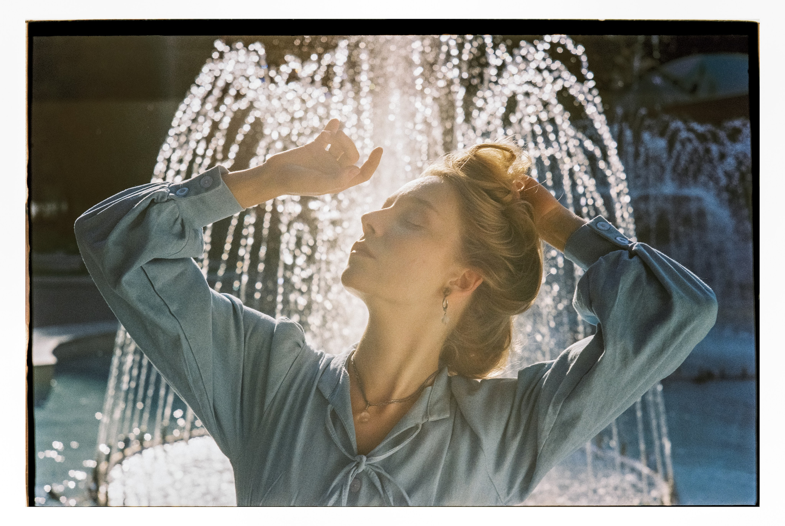 Портрет девушки со светлыми волосами в синем платье в контровом солнечном свете на фоне фонтана