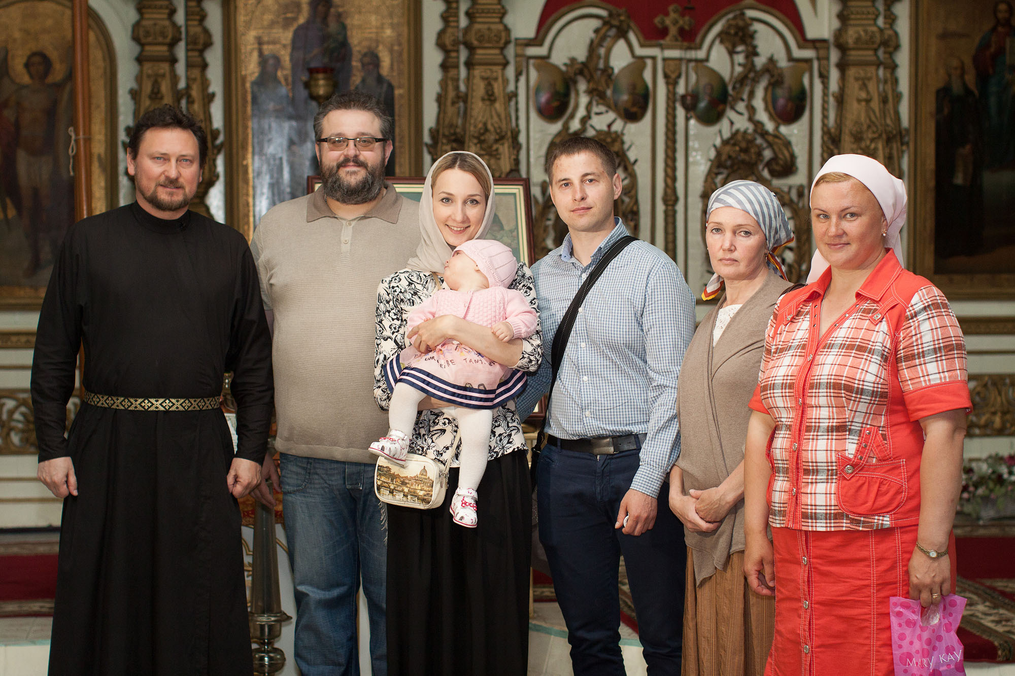 Родственники на крестинах стоят в 1 шеренгу со священником в церкви.